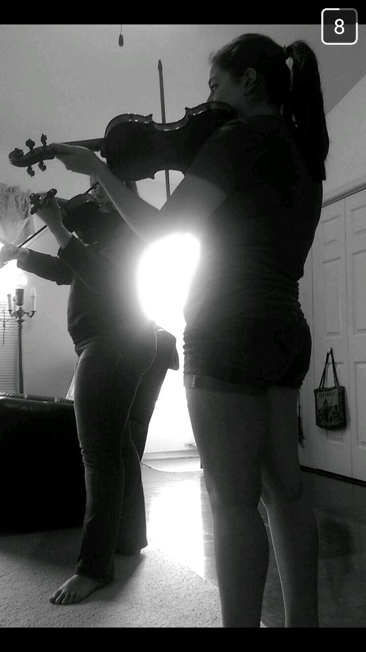 Two women playing guitar indoors, one facing sideways, with a bright light behind them, in a room with a cabinet and a bag.