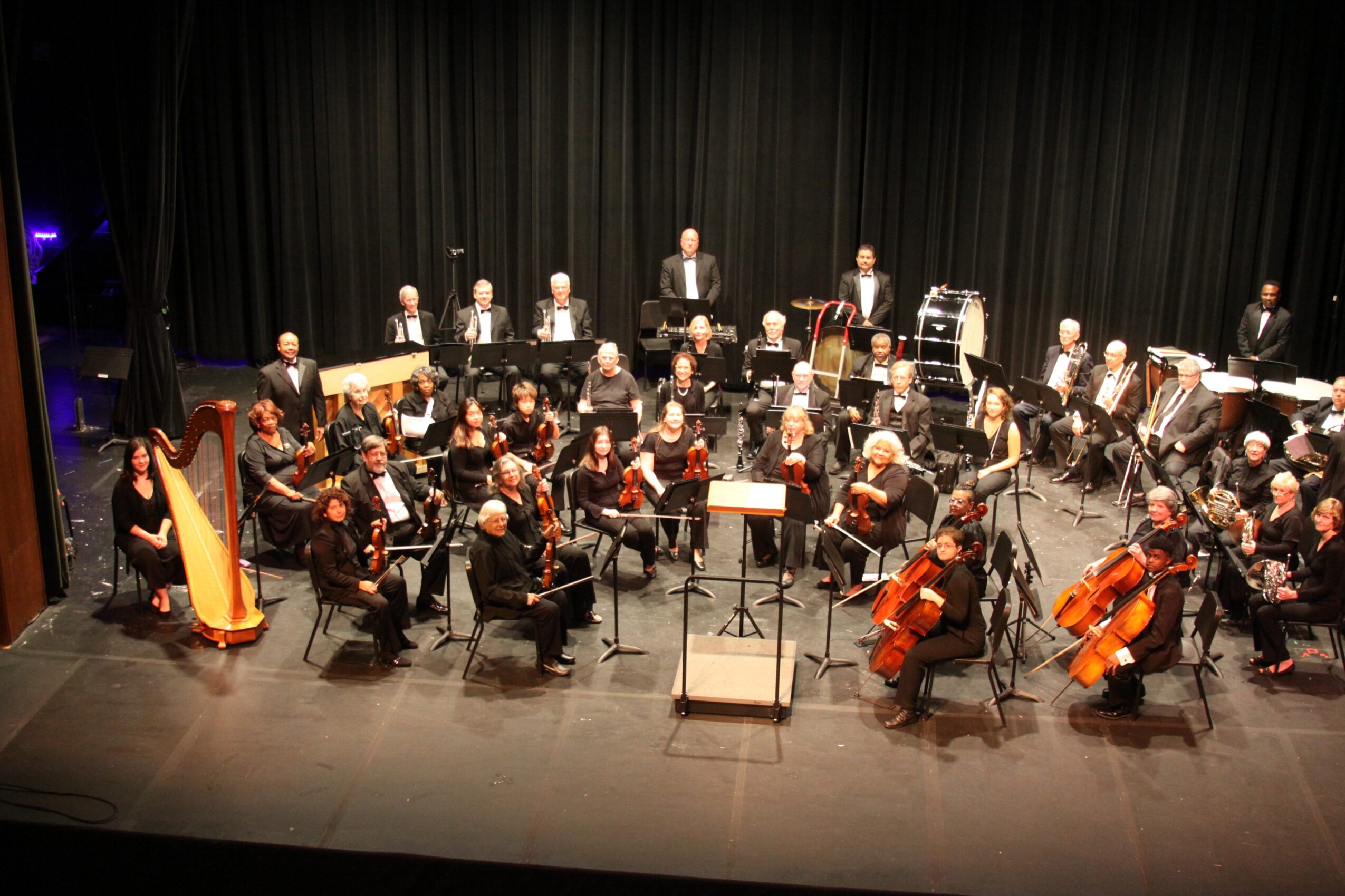 Orchestra performing on stage with musicians playing various instruments, conductor in front, black curtain backdrop, some percussion instruments visible.