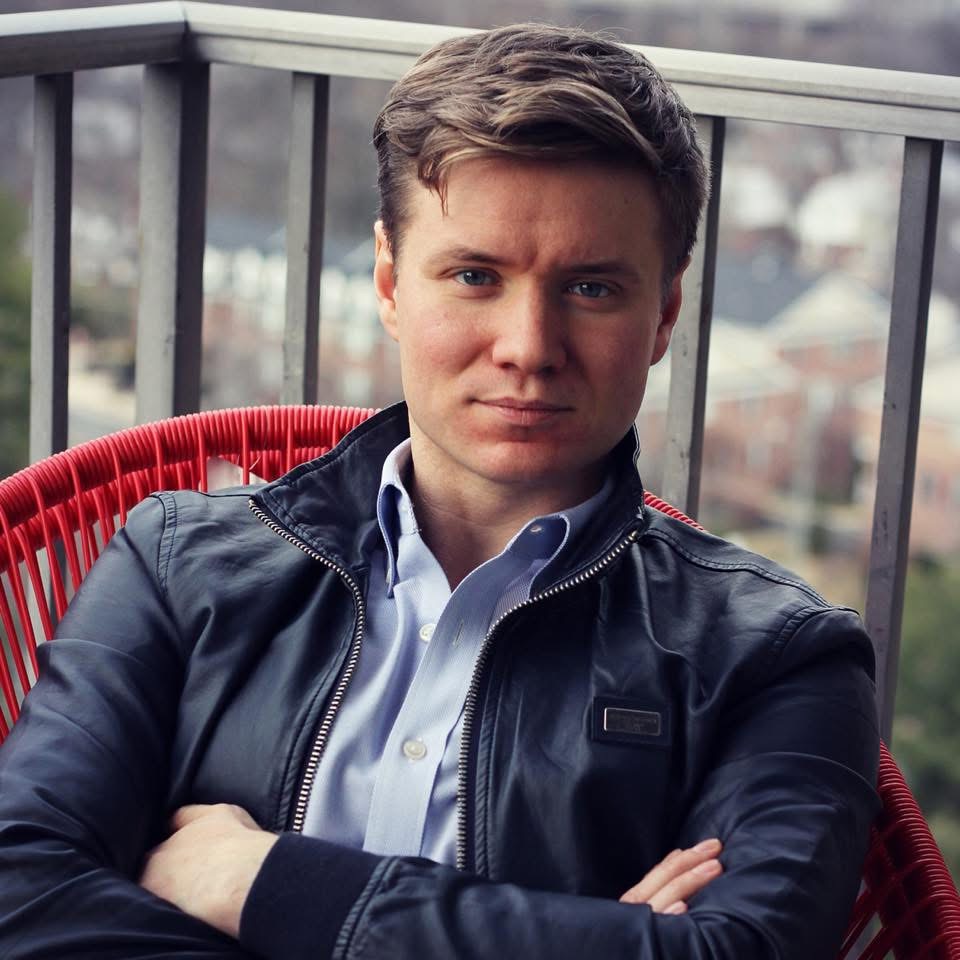 Young man with short brown hair, wearing a black leather jacket and light blue shirt, sitting outdoors with arms crossed.