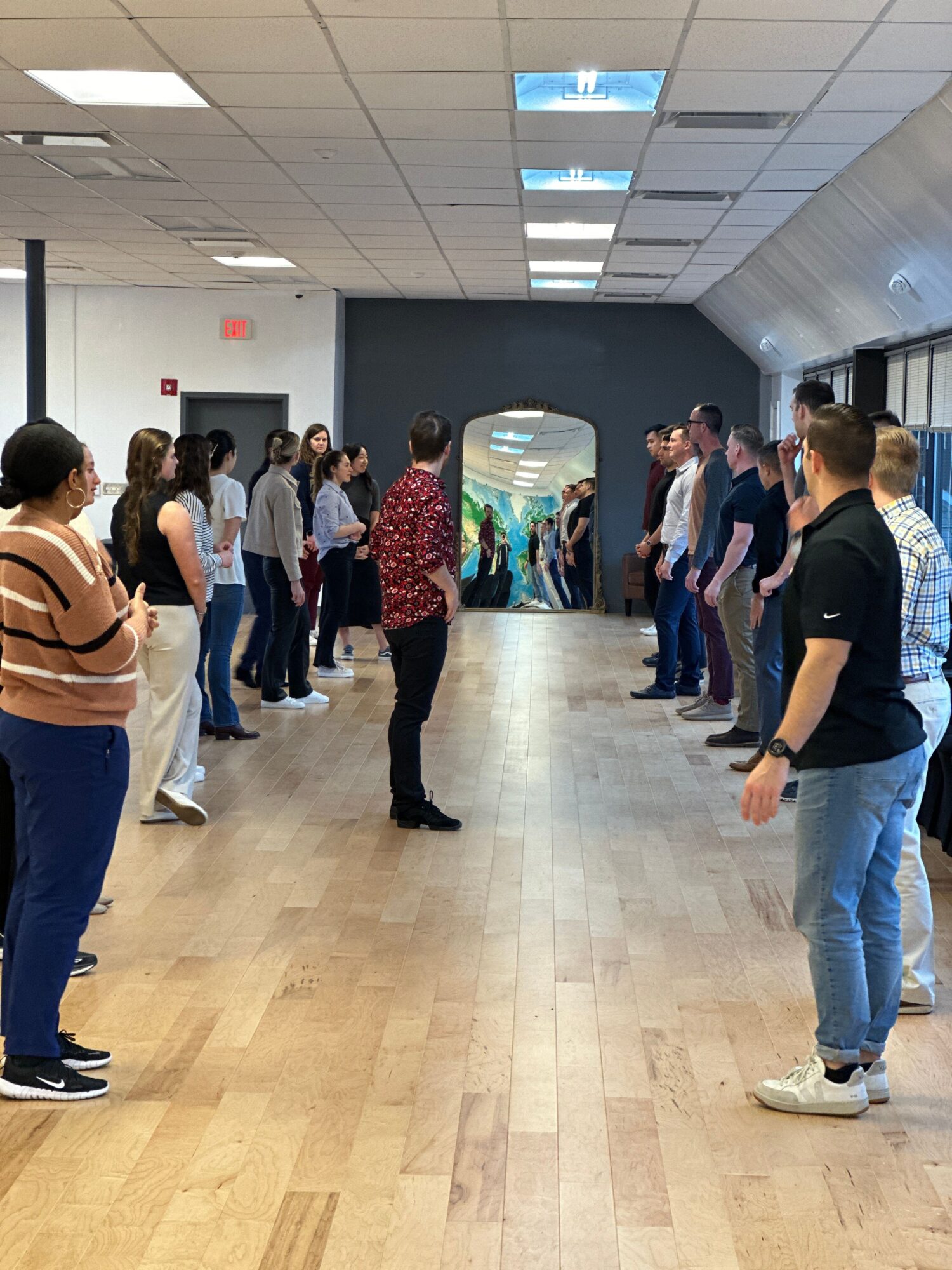 Group of people standing in line in a hallway with a curved wall and wooden floor, some looking at each other.
