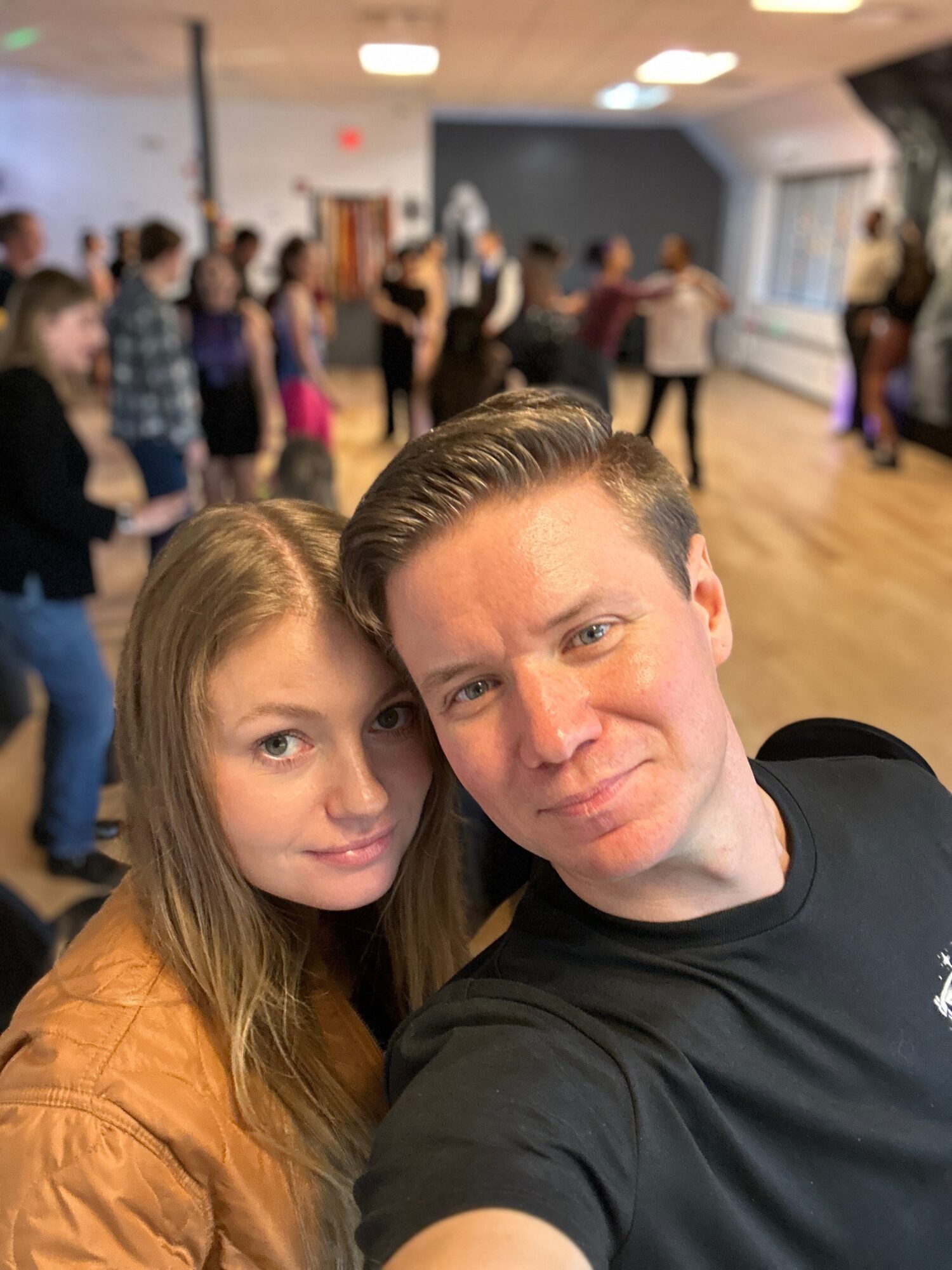 Two young people taking a selfie in a room with a group of people dancing in the background.