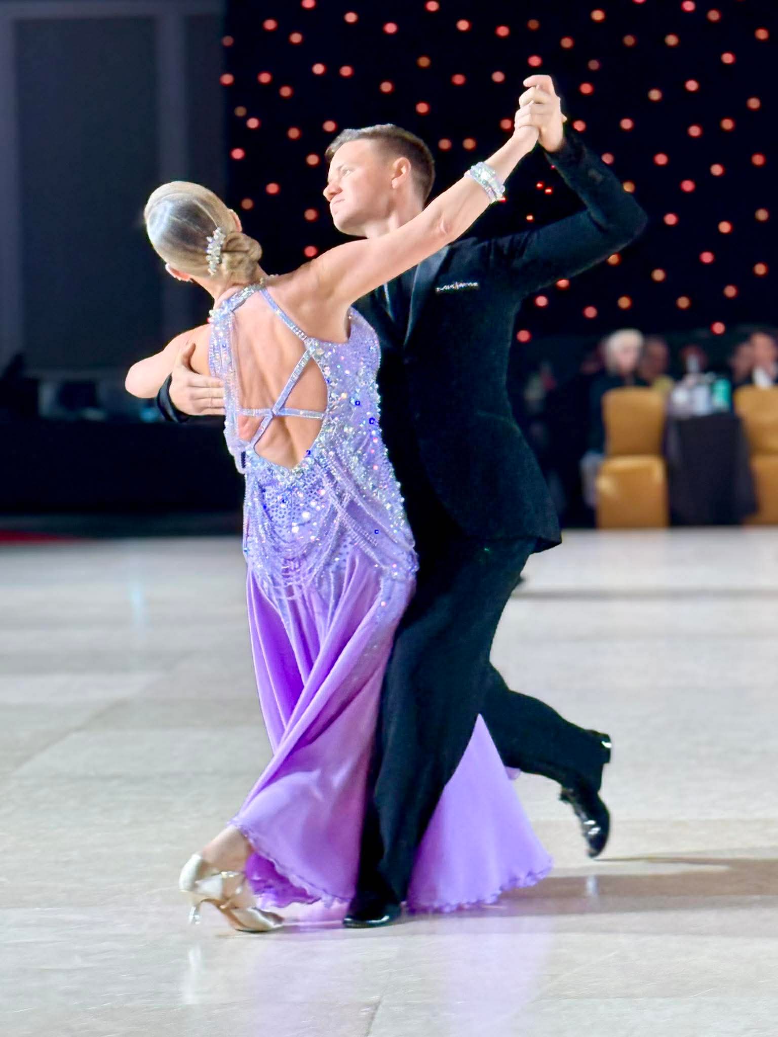 A couple dances on a ballroom floor, the woman in a purple dress and the man in a black suit, holding hands.