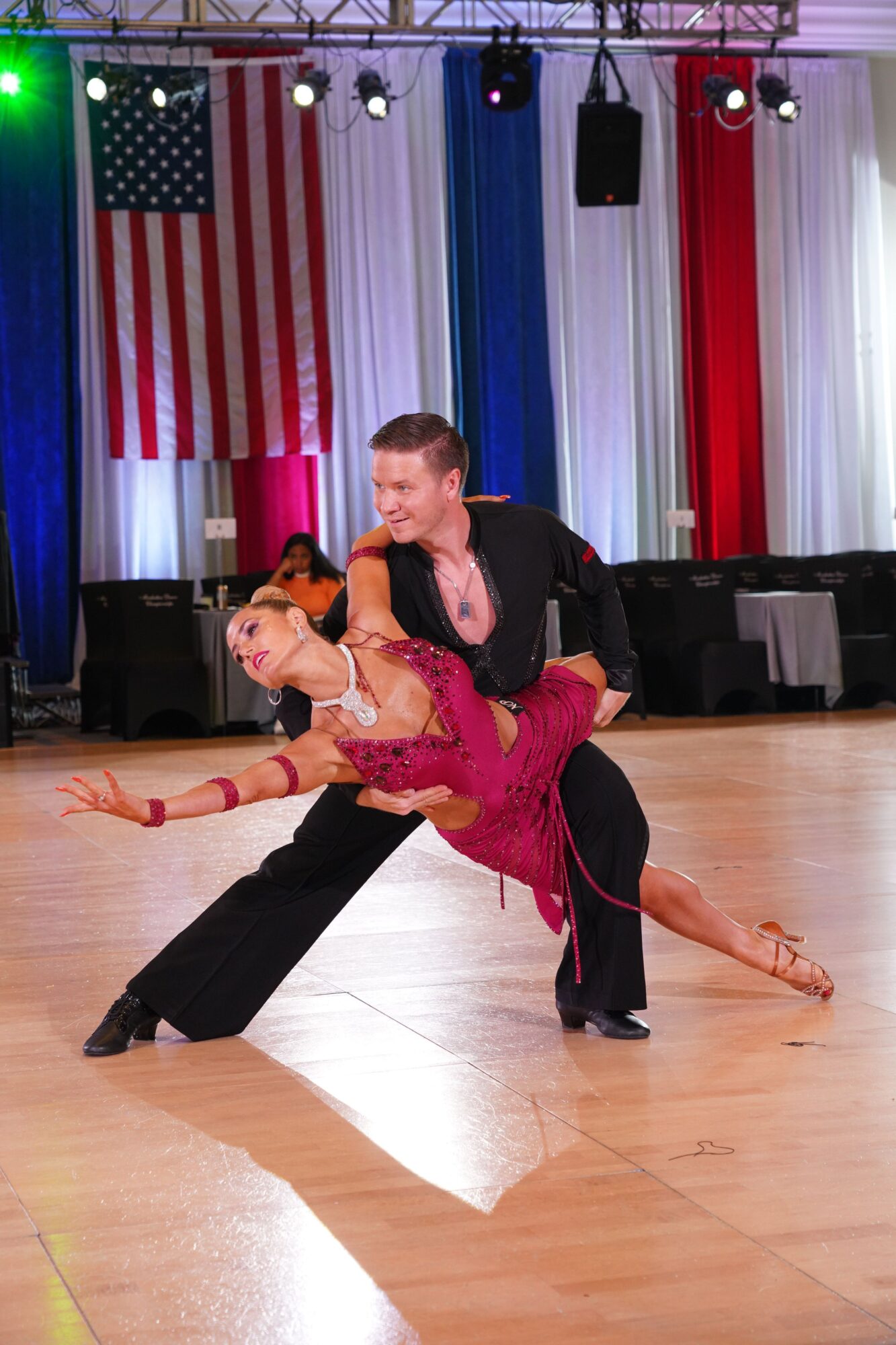 A couple dancing on a wooden floor with an American flag backdrop, stage lights, and speakers in the background.