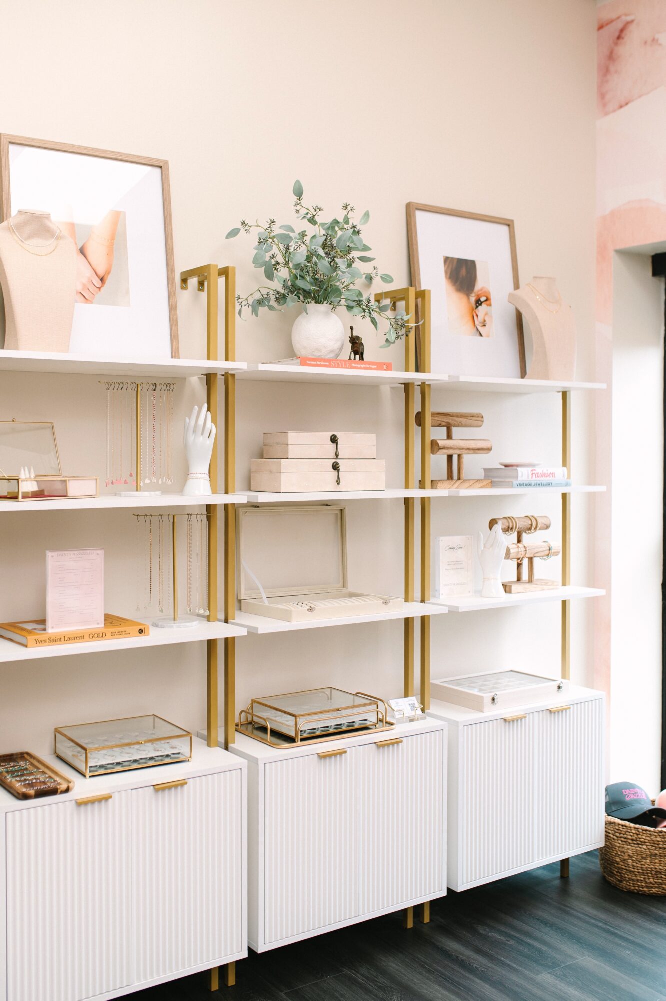 White shelving unit with decorative items, books, and framed pictures, against a light-colored wall with pink accents, on a dark floor.
