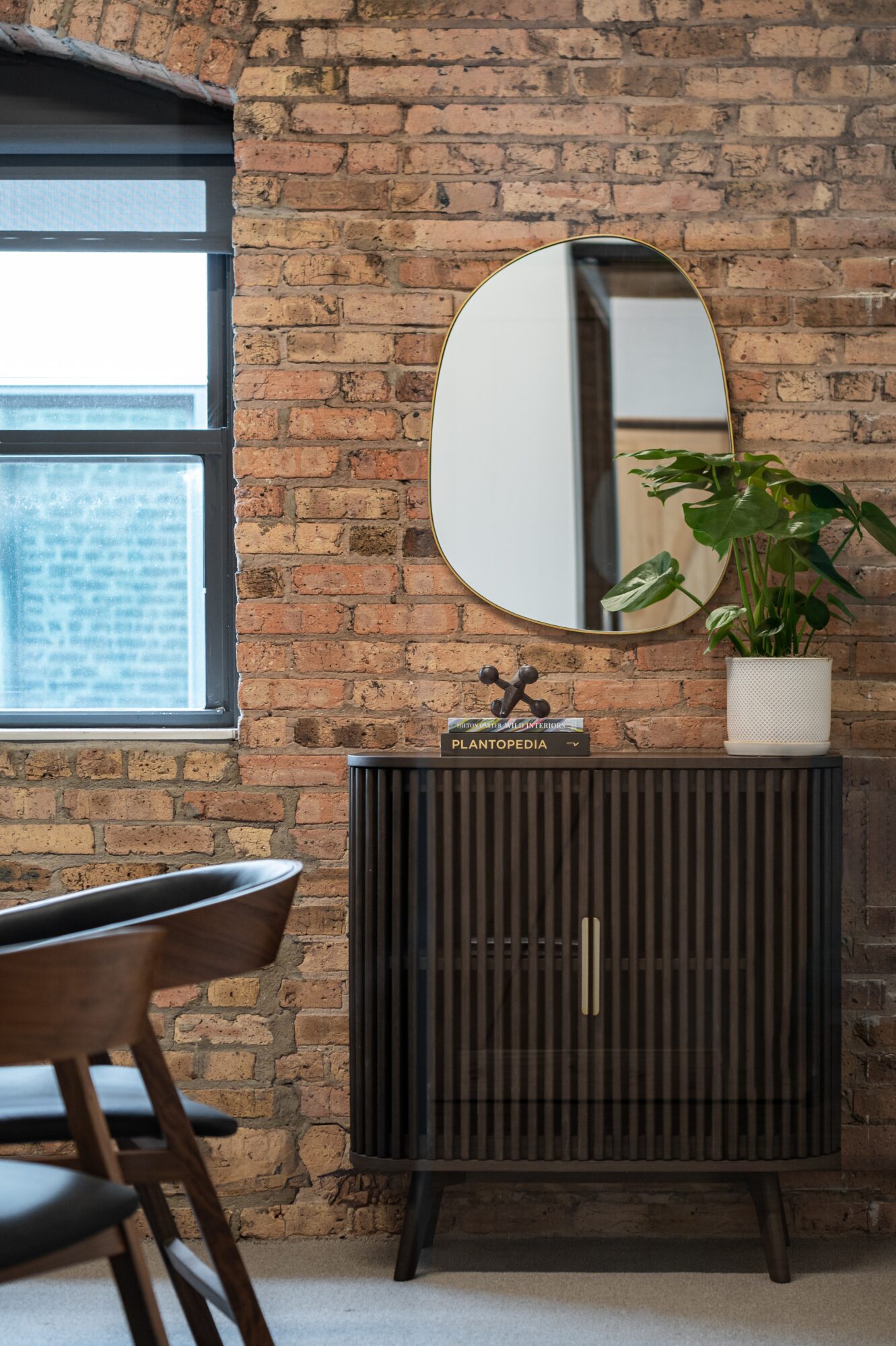 Interior scene with a brick wall, round mirror, window, and a black cabinet with a plant on top.