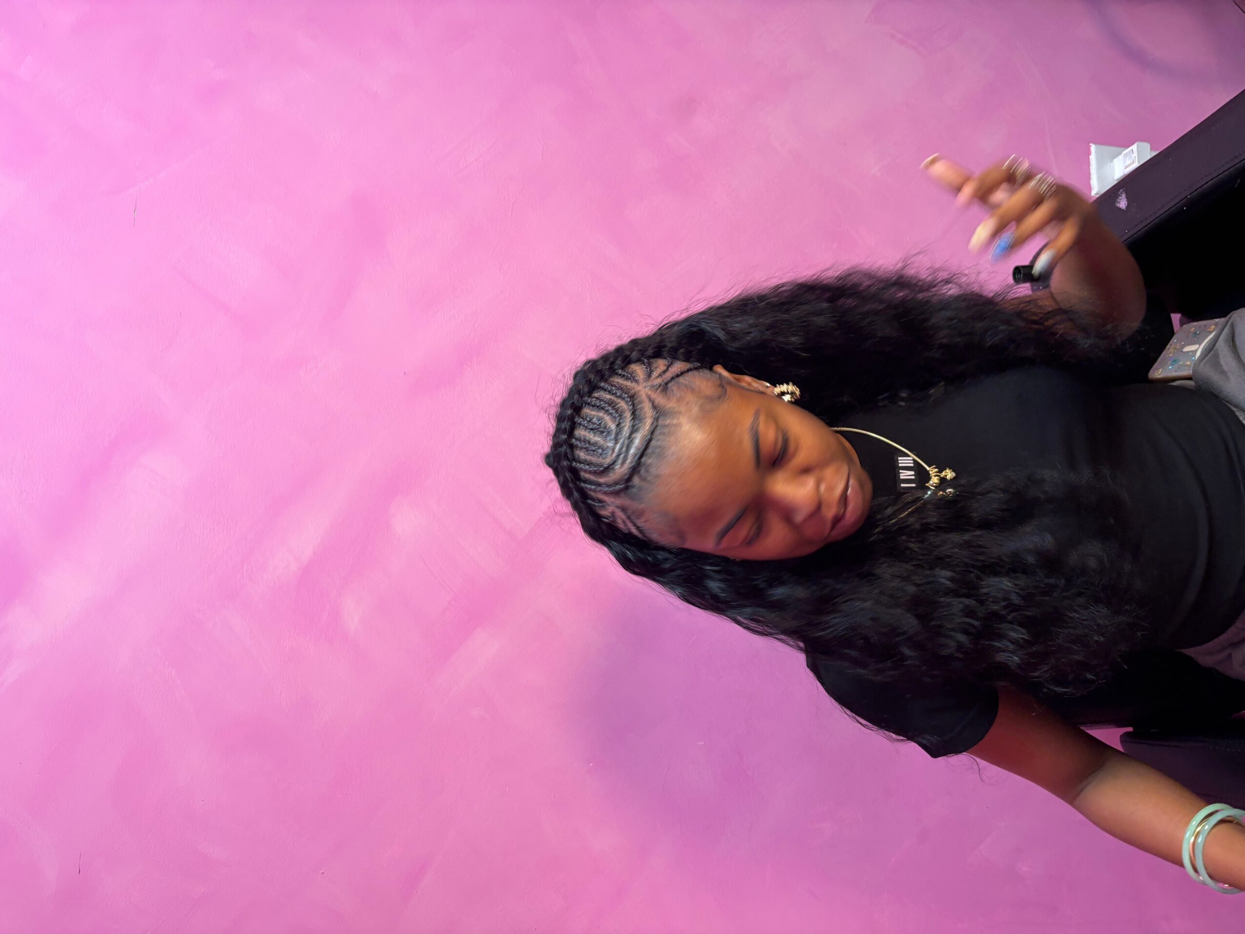 Woman with braided hair and jewelry, smiling, against a pink wall, making a peace sign with her hand.