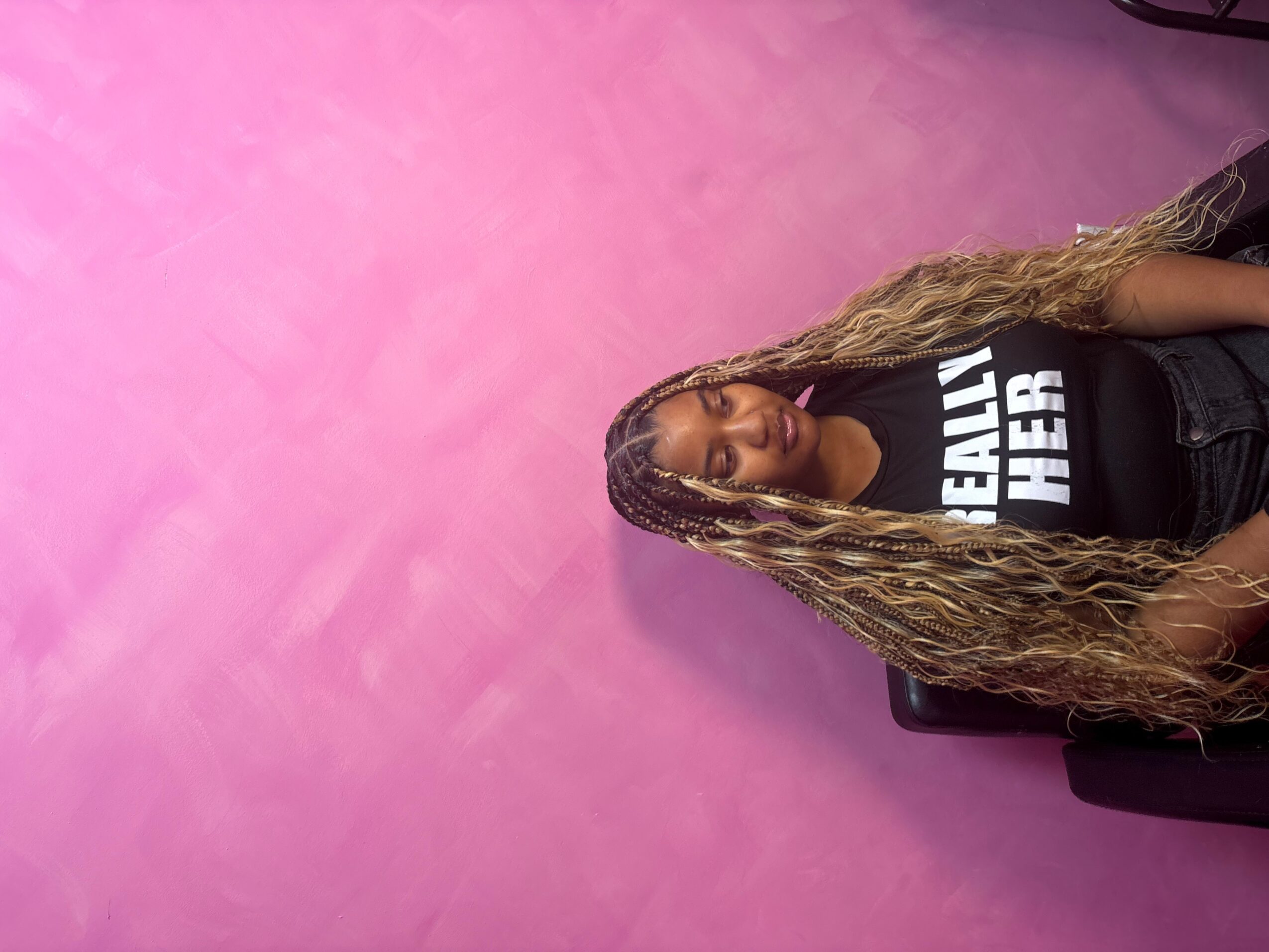 Woman with long curly hair sitting against a pink wall, wearing a black T-shirt with white text.