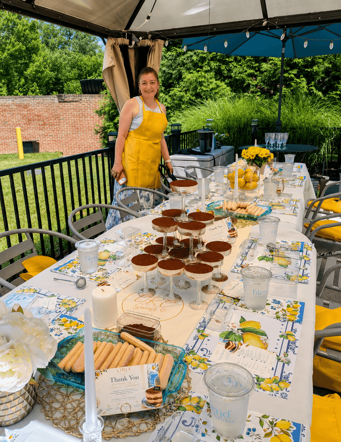 Woman in yellow apron standing behind a decorated table outdoors, with desserts and drinks, under a canopy, greenery in background.