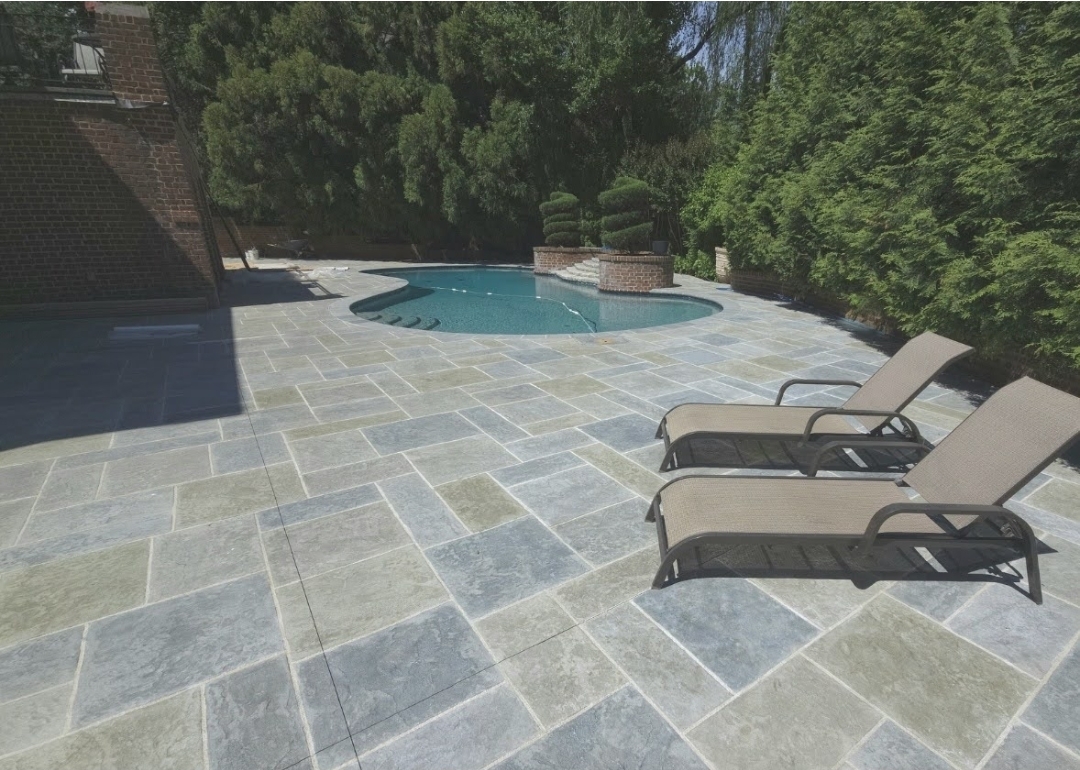 Swimming pool with two lounge chairs on a tiled patio, surrounded by trees and greenery.