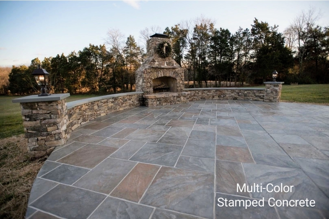 Outdoor stone fireplace with a curved stone patio and a low stone wall, surrounded by trees and grass.