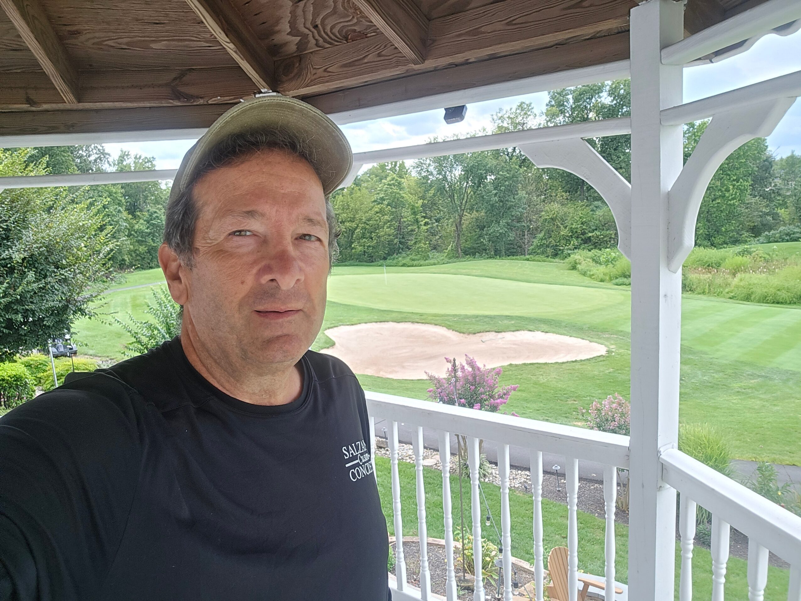 Man wearing a cap and black shirt standing on a porch overlooking a golf course with trees and sand traps.