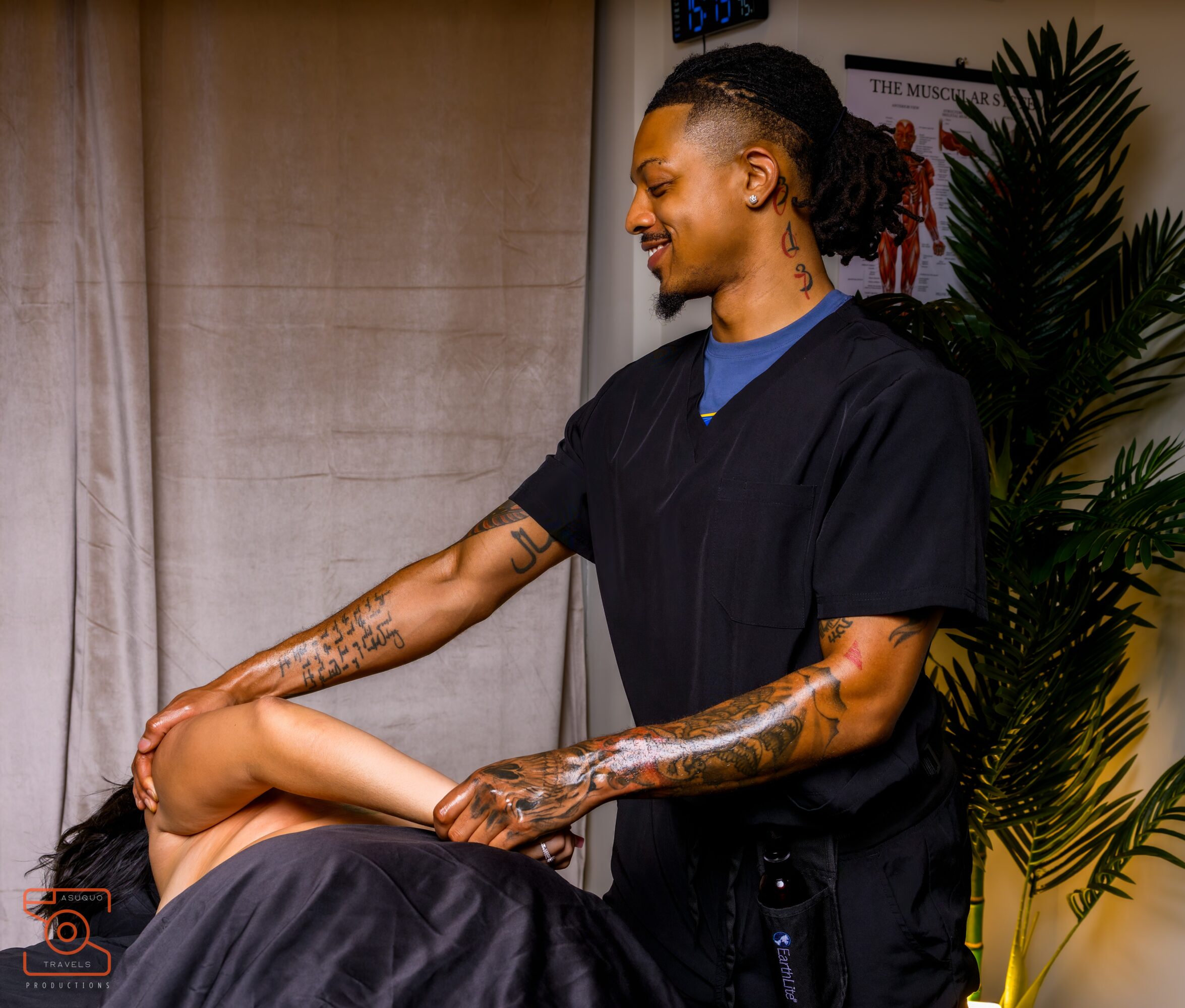 Person receiving a massage from a therapist with tattoos, smiling, in a room with a curtain and plant.