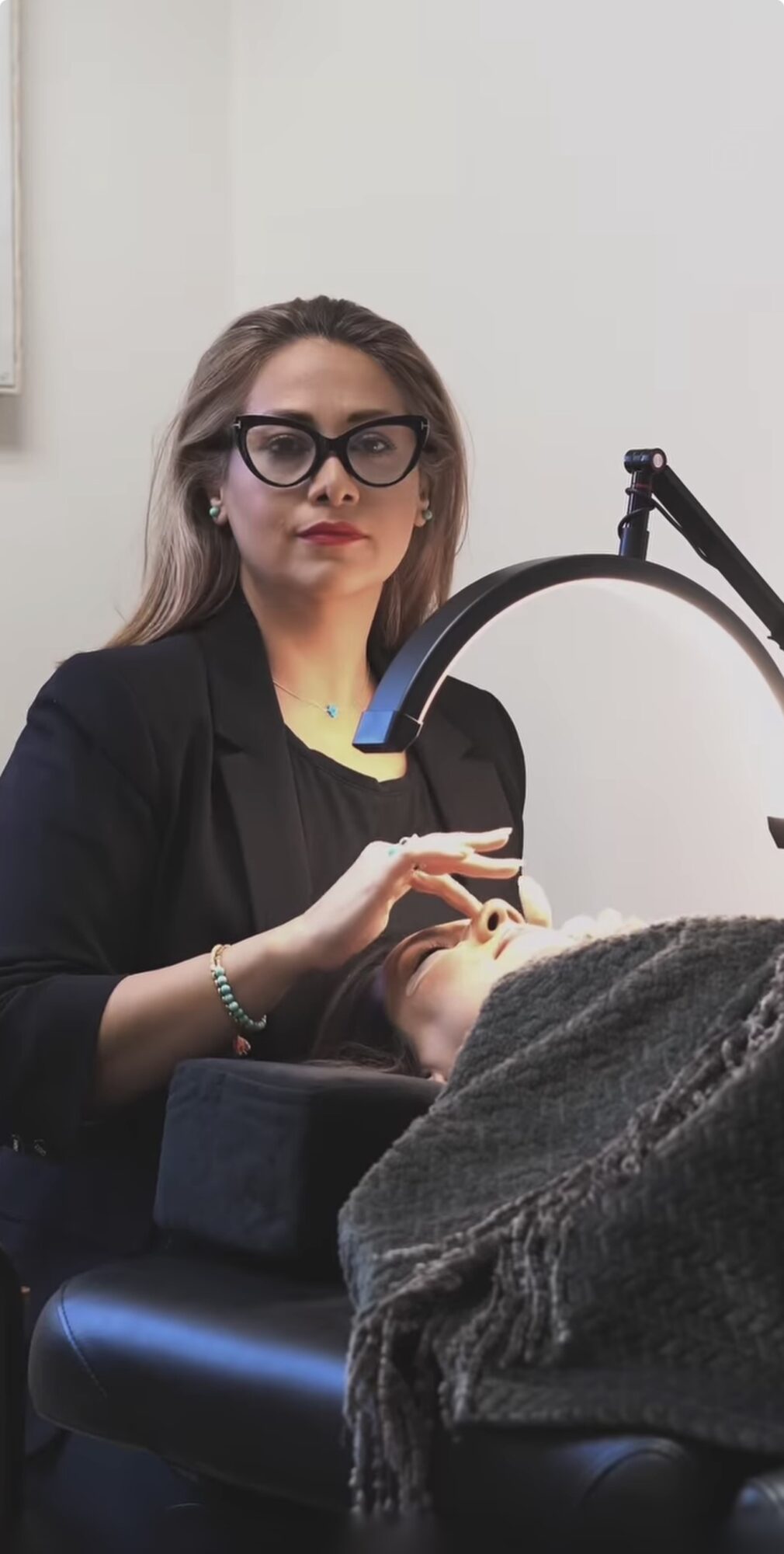 Woman with glasses performs a procedure on a person's face in a clinical setting.