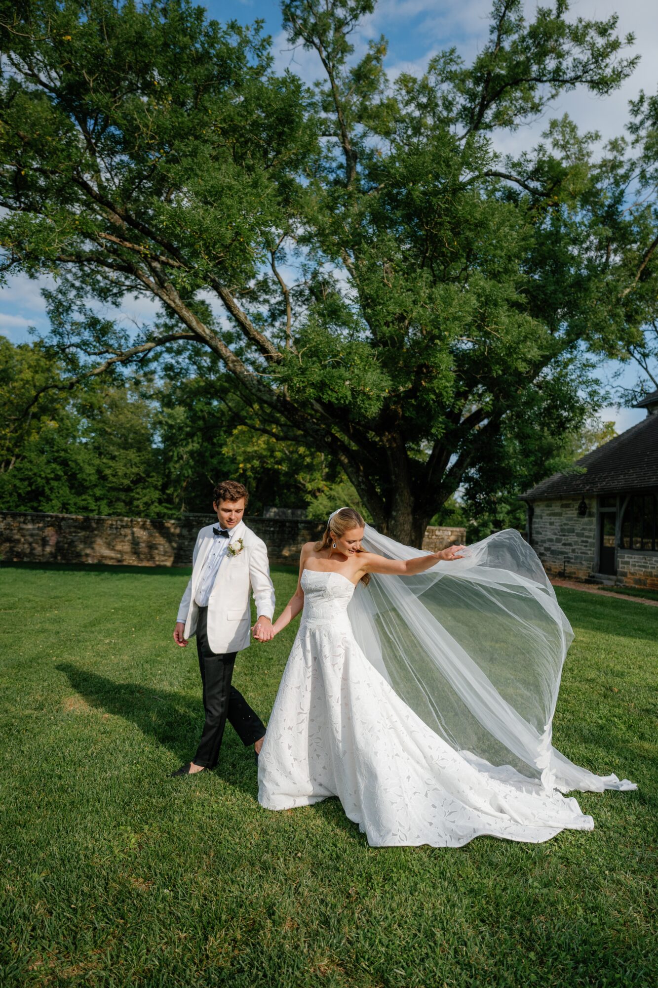 Bride and groom holding hands outdoors with a large tree in the background, bride in a white dress and veil, groom in a suit.