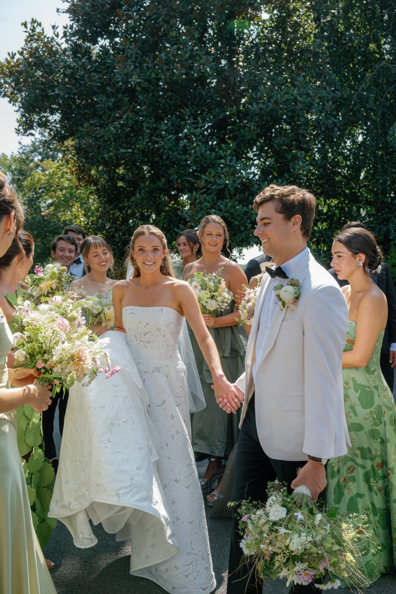 A wedding ceremony outdoors with a bride and groom holding hands, surrounded by bridesmaids and guests, trees in background.