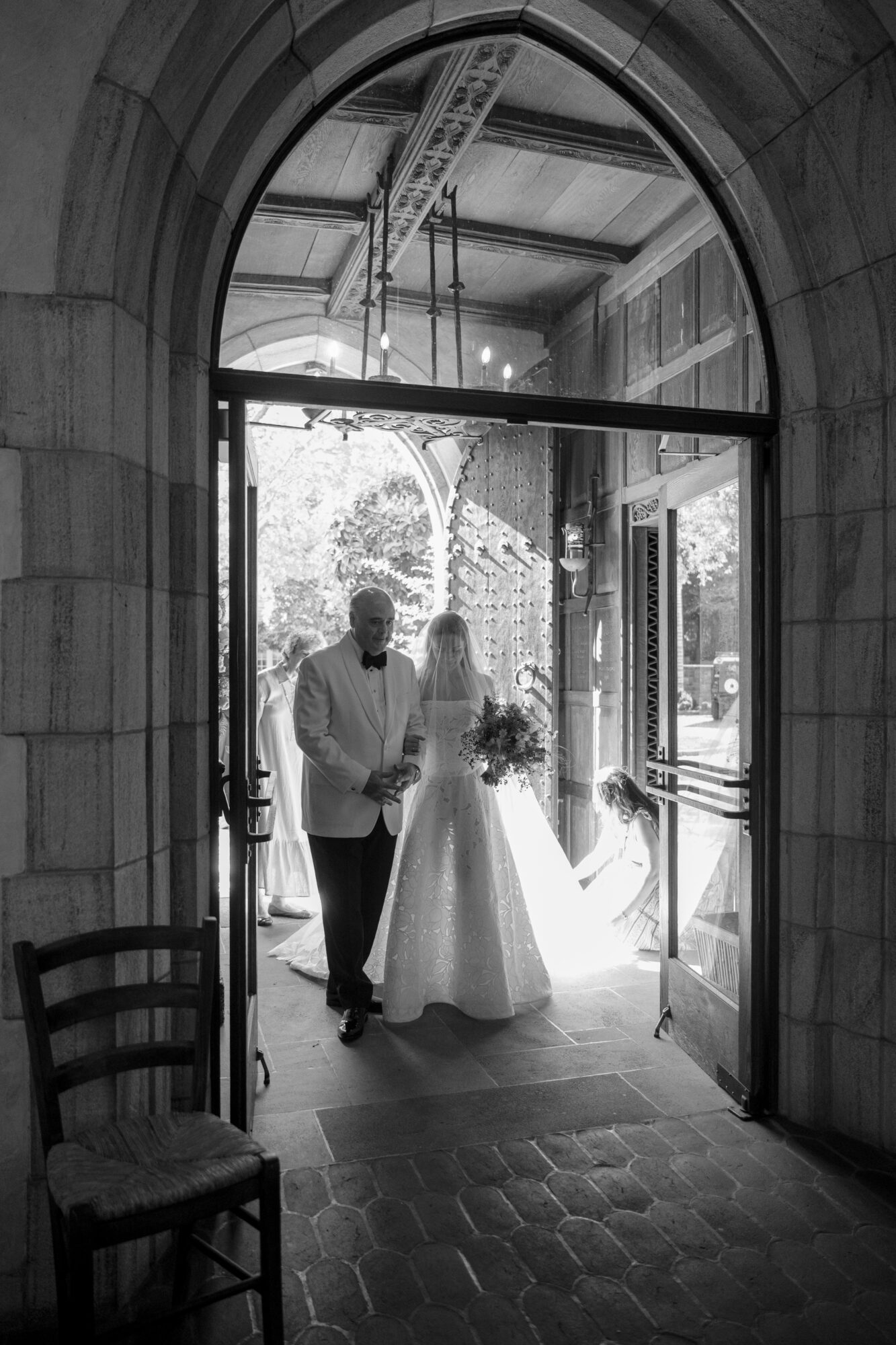 A person and a bride holding a bouquet walk through an arched doorway into a bright room, with sunlight and trees outside.