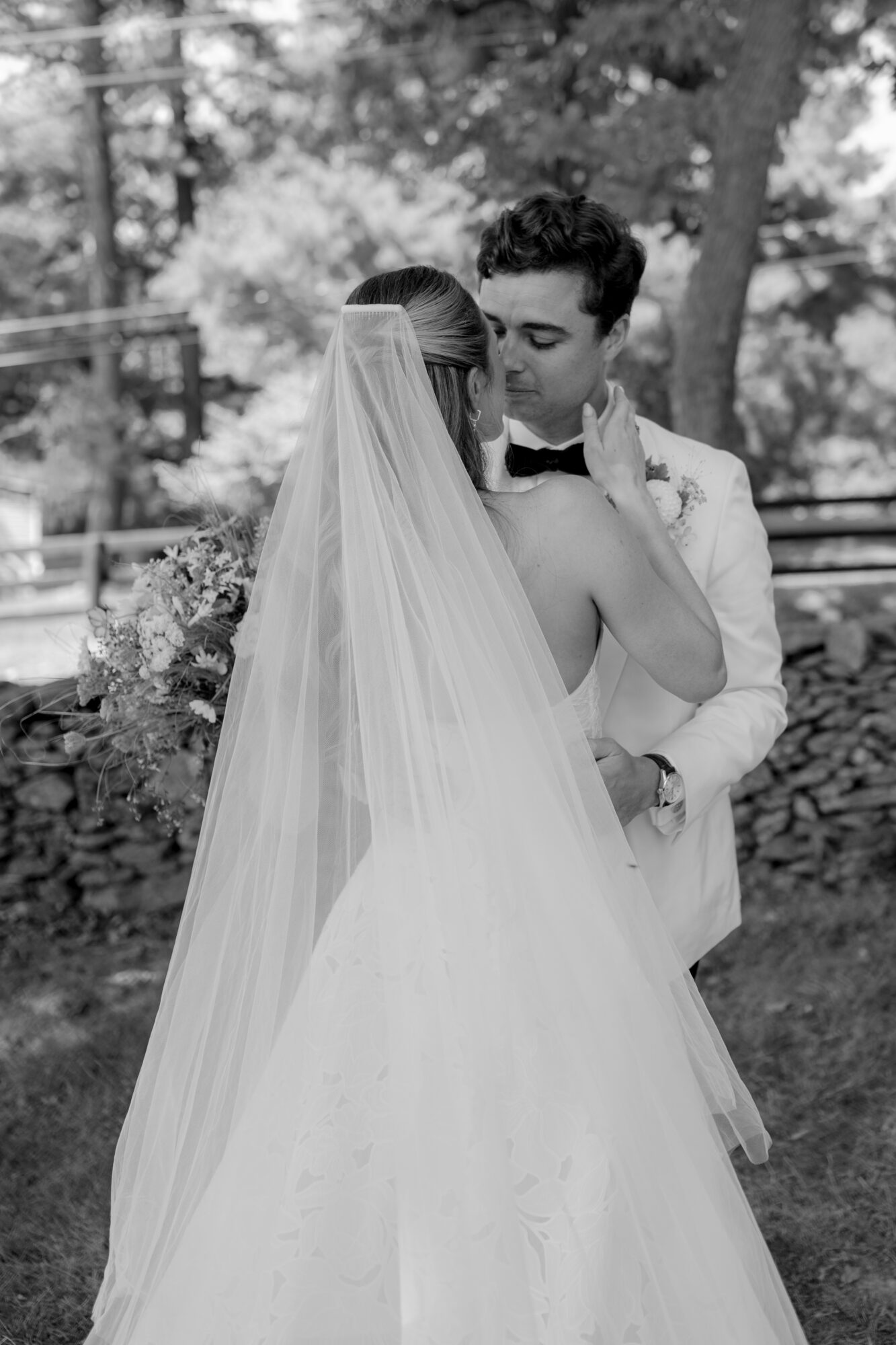 Bride and groom embrace outdoors, bride in wedding dress with veil, groom in tuxedo, trees in background