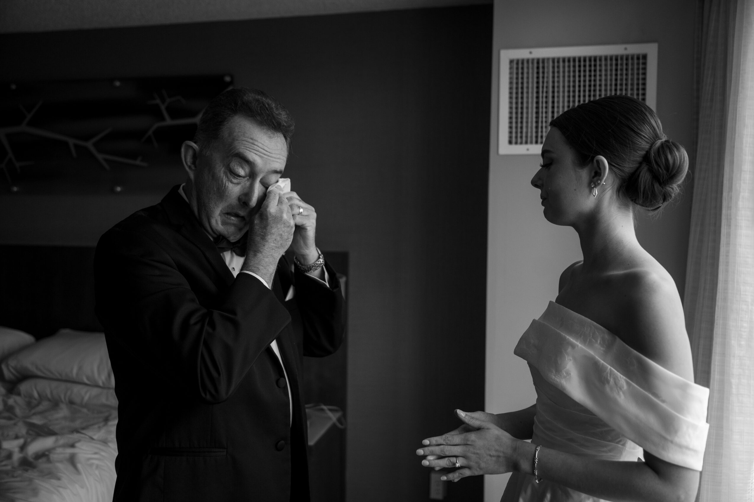 A man with glasses and a woman in an off-shoulder dress face each other indoors, with the man appearing emotional.