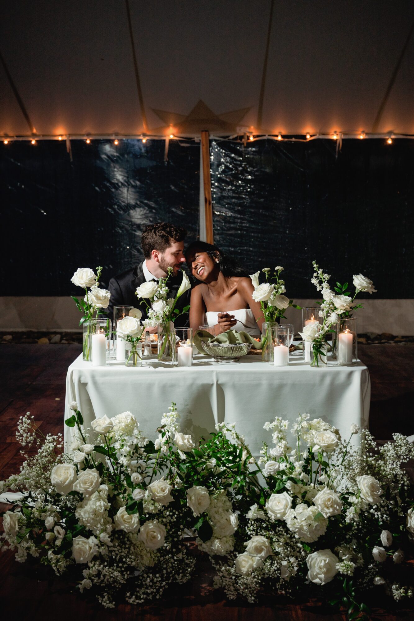 Couple kissing at a decorated table with white flowers, candles, and a dark water background, under a tent at night.