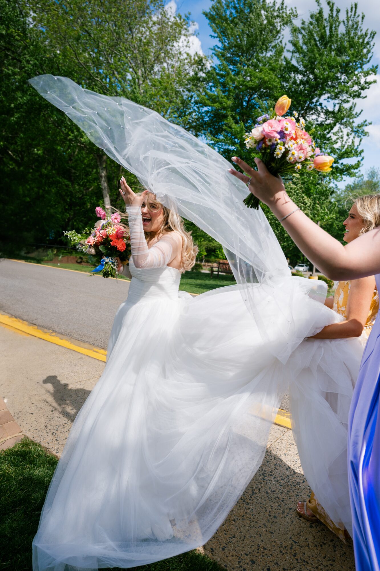 Bride in wedding dress holding bouquet, veil lifted by another person outdoors with trees and sky.