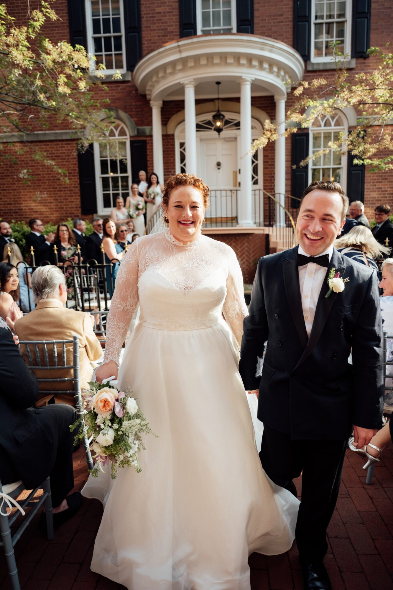 Bride and groom smiling outdoors in front of a historic building with columns, wedding guests seated around them.