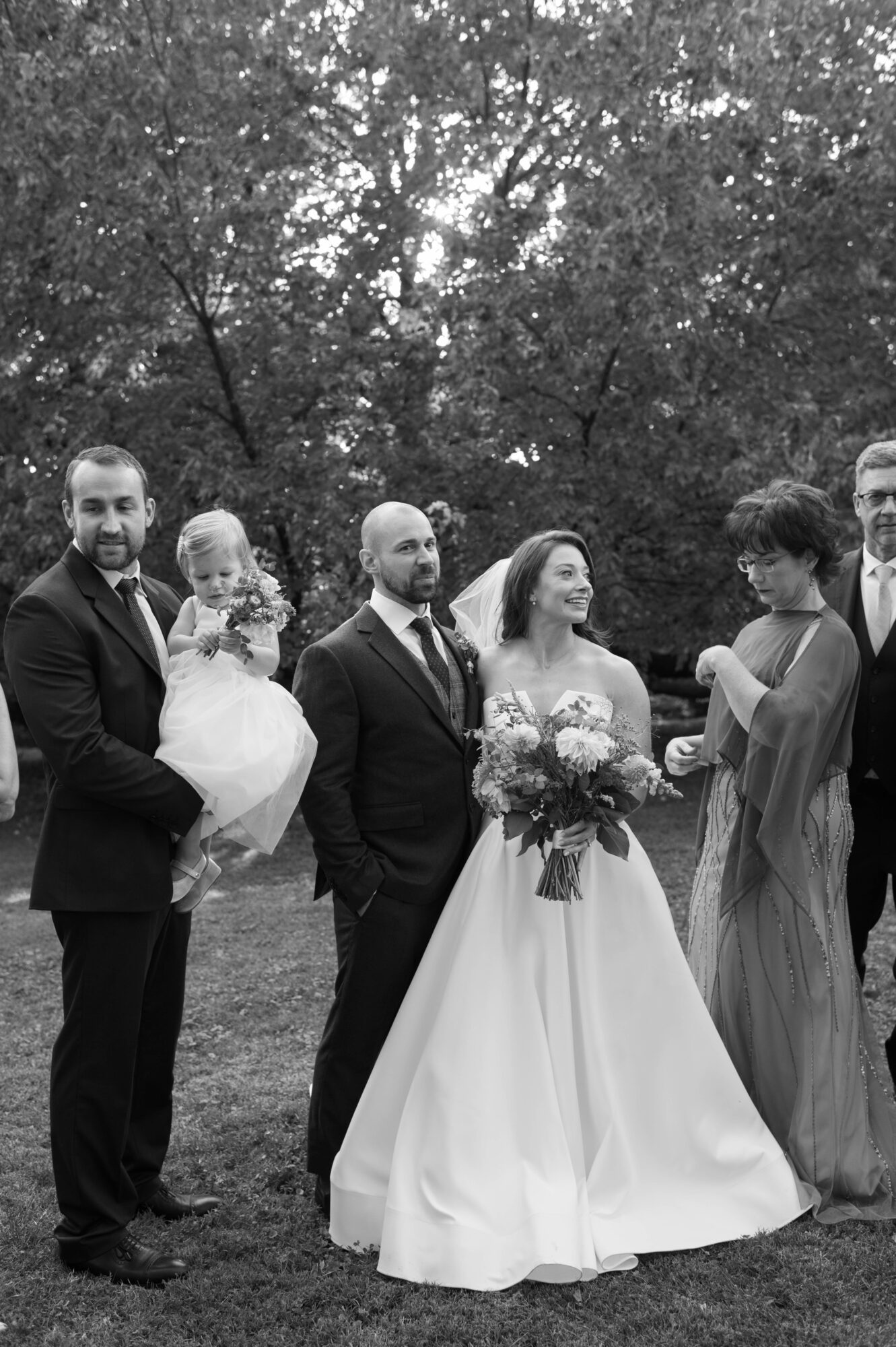 Group of six people outdoors, including a bride holding a bouquet, and others in formal attire, trees in background.