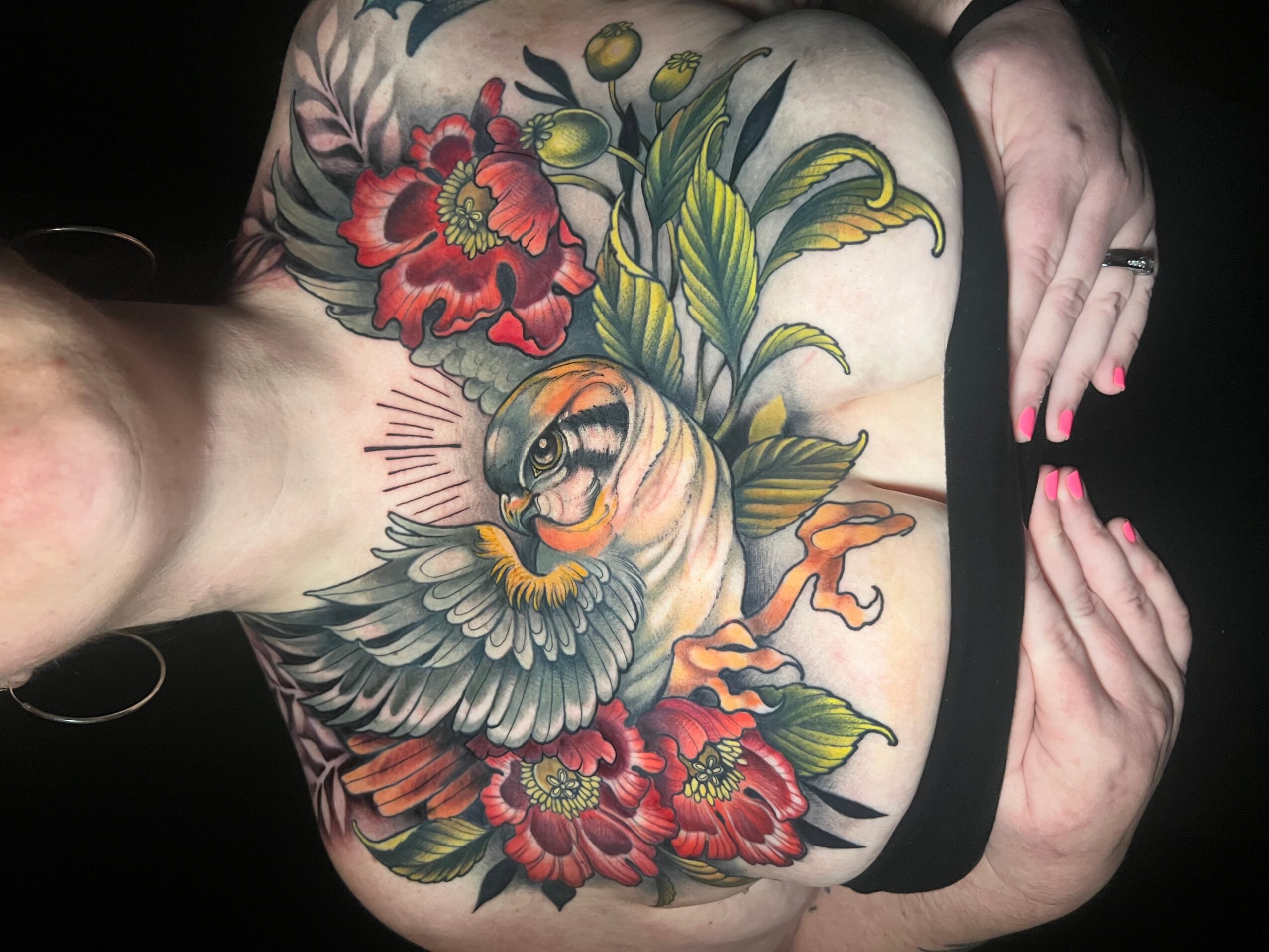 Colorful tattoo of a bird surrounded by flowers on person's shoulder and back, with hand resting nearby.