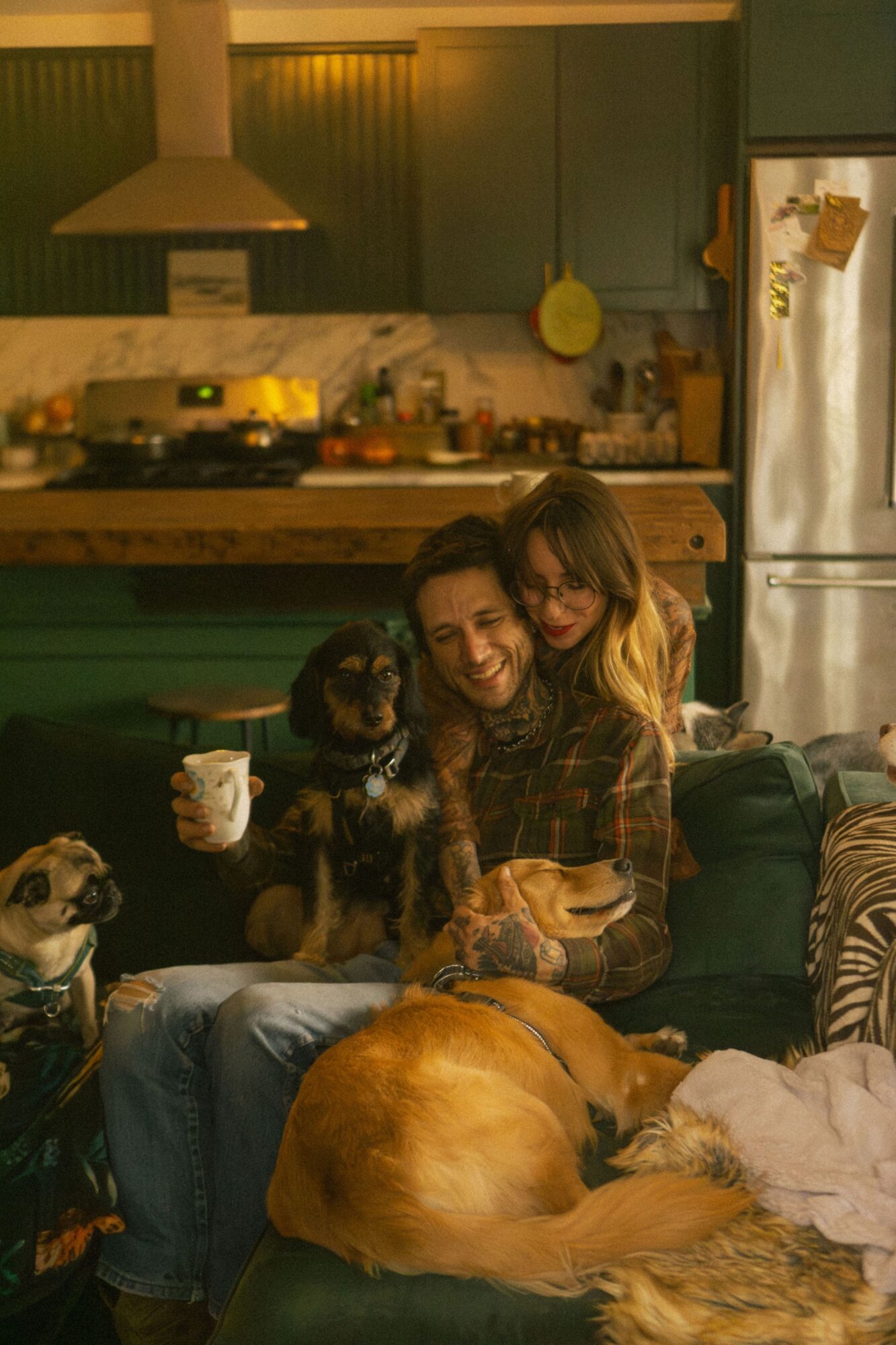 Person sitting on a couch with three dogs, holding a mug, in a kitchen and living room setting.