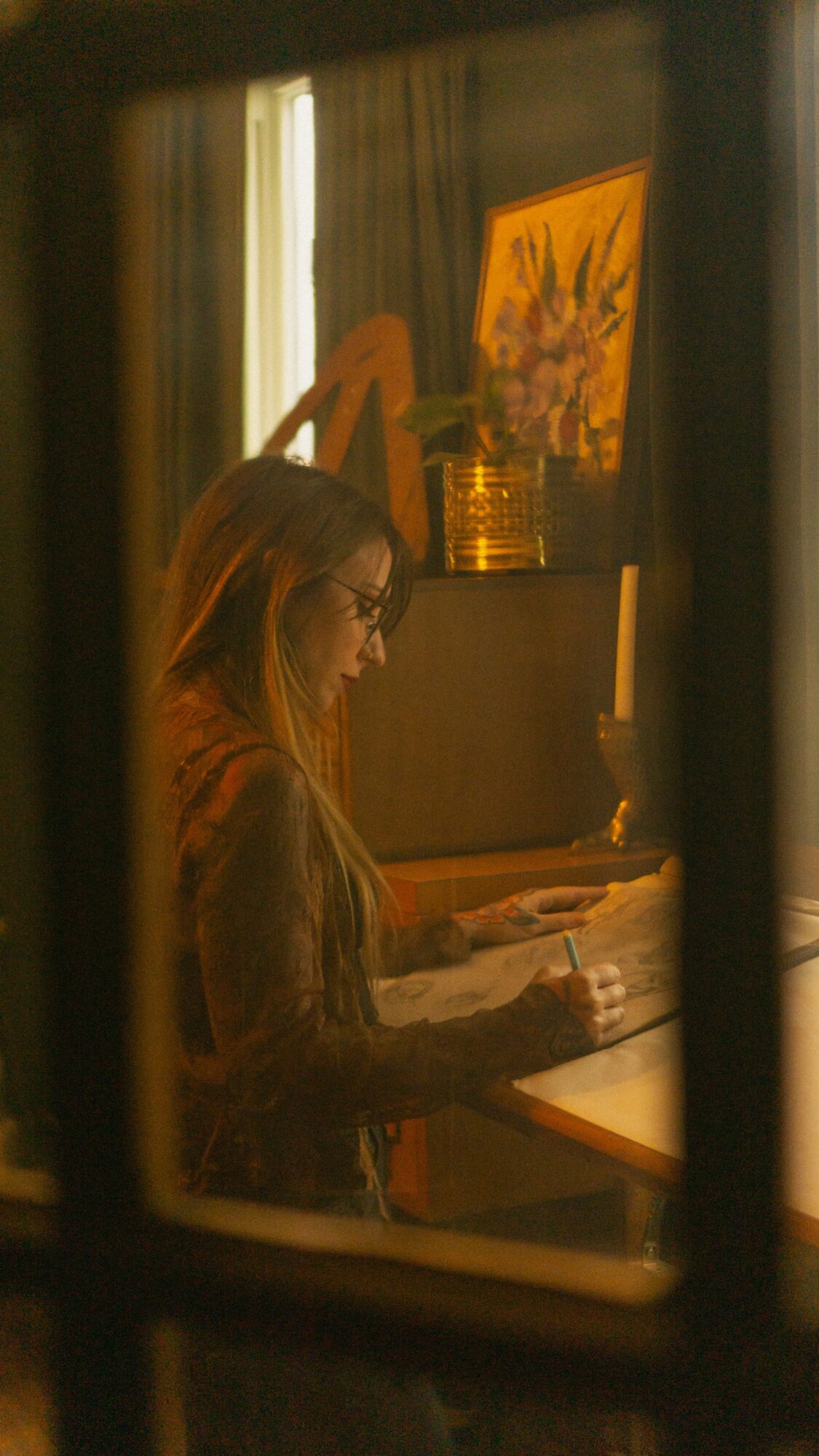 Woman with glasses reading a book at a table, warm indoor lighting, framed by window panes.