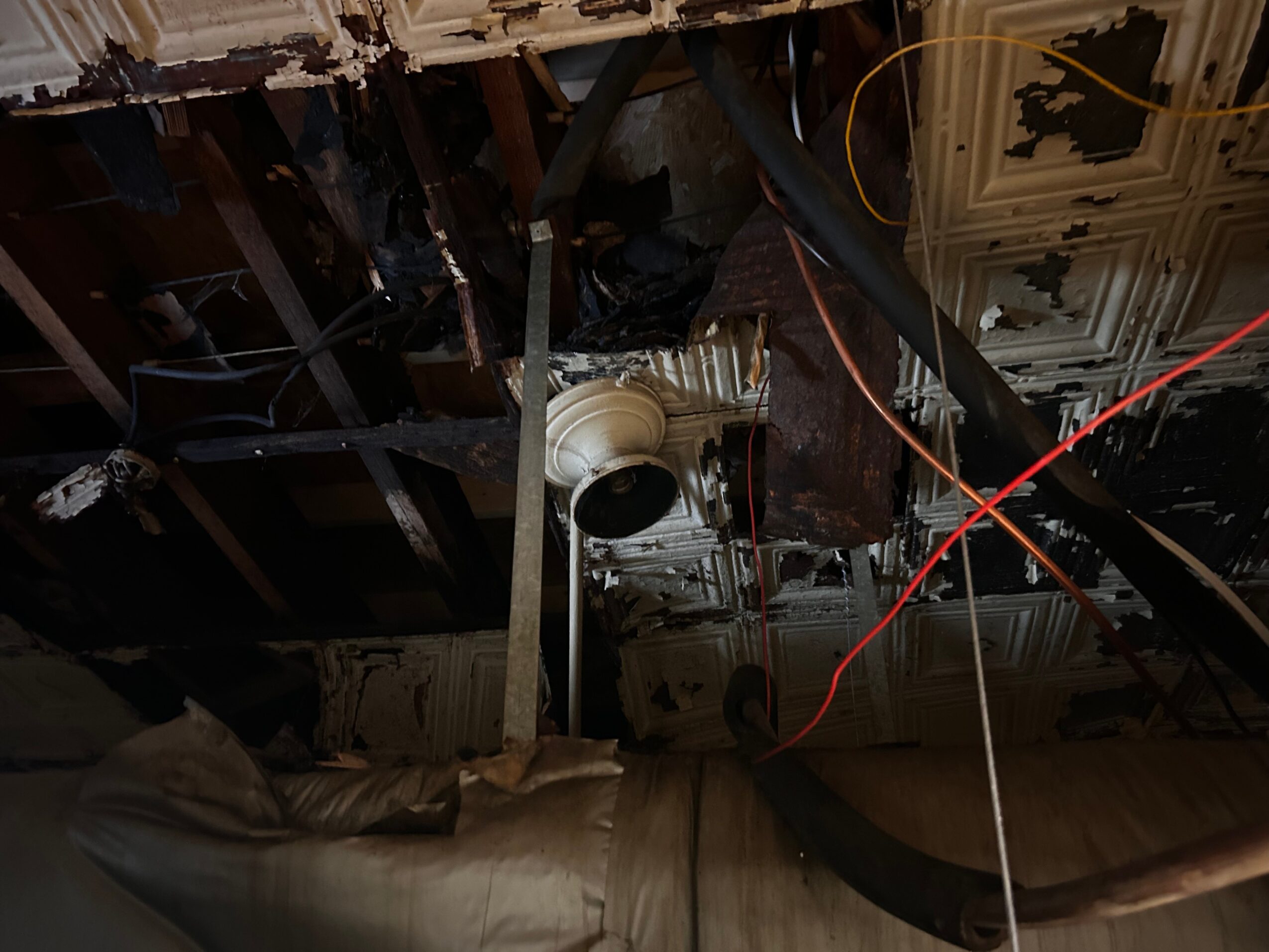 Ceiling with exposed wooden beams, damaged insulation, and hanging wires, with a partially visible ceiling vent and damaged ceiling tiles.