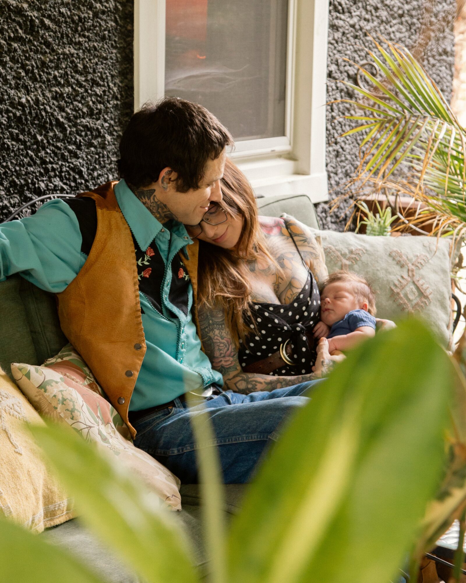 Two adults and a newborn baby sitting on a sofa outdoors, with plants and a window behind them.