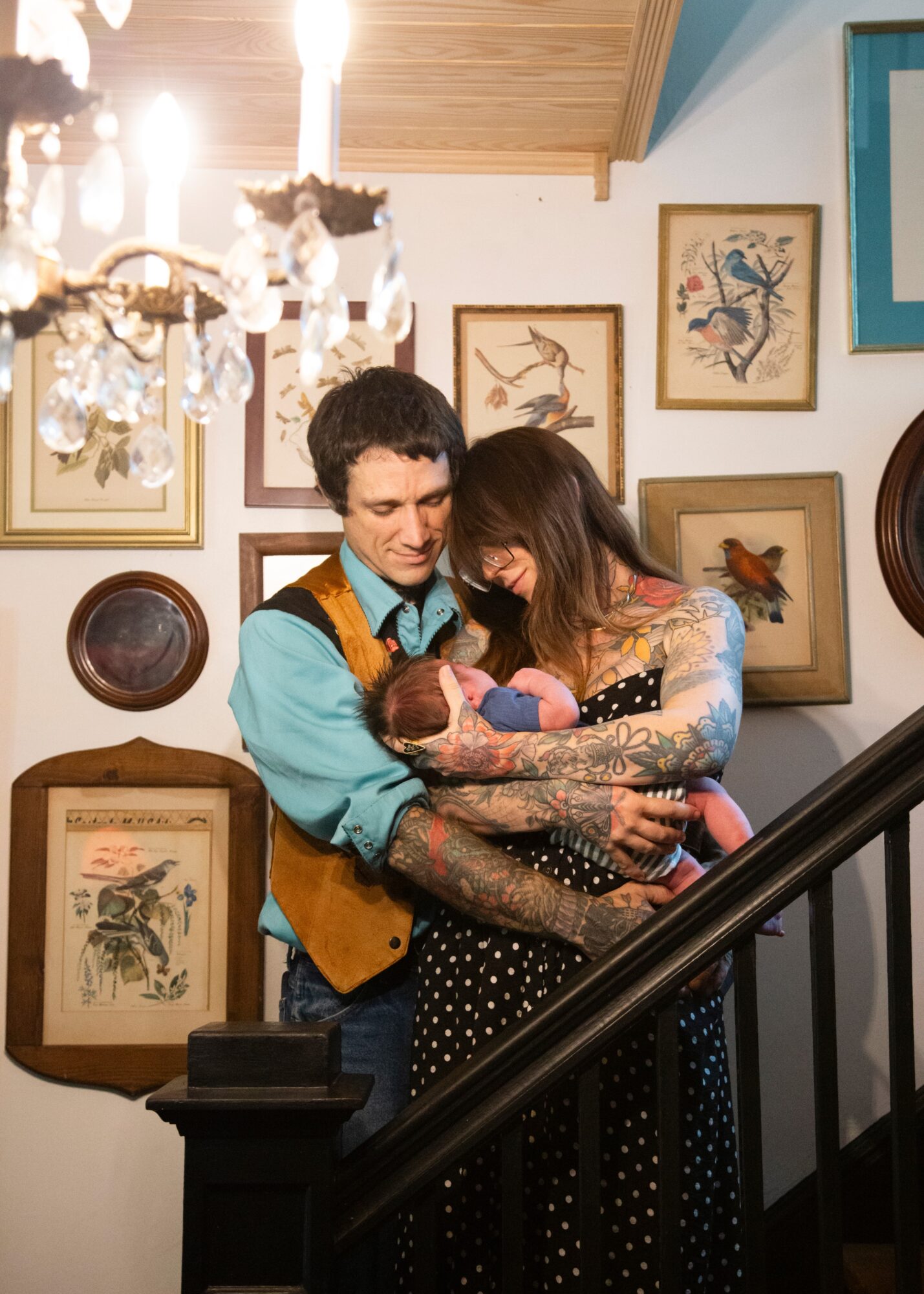 Two women holding a small dog on a staircase, surrounded by framed bird artwork on the wall, chandelier overhead.