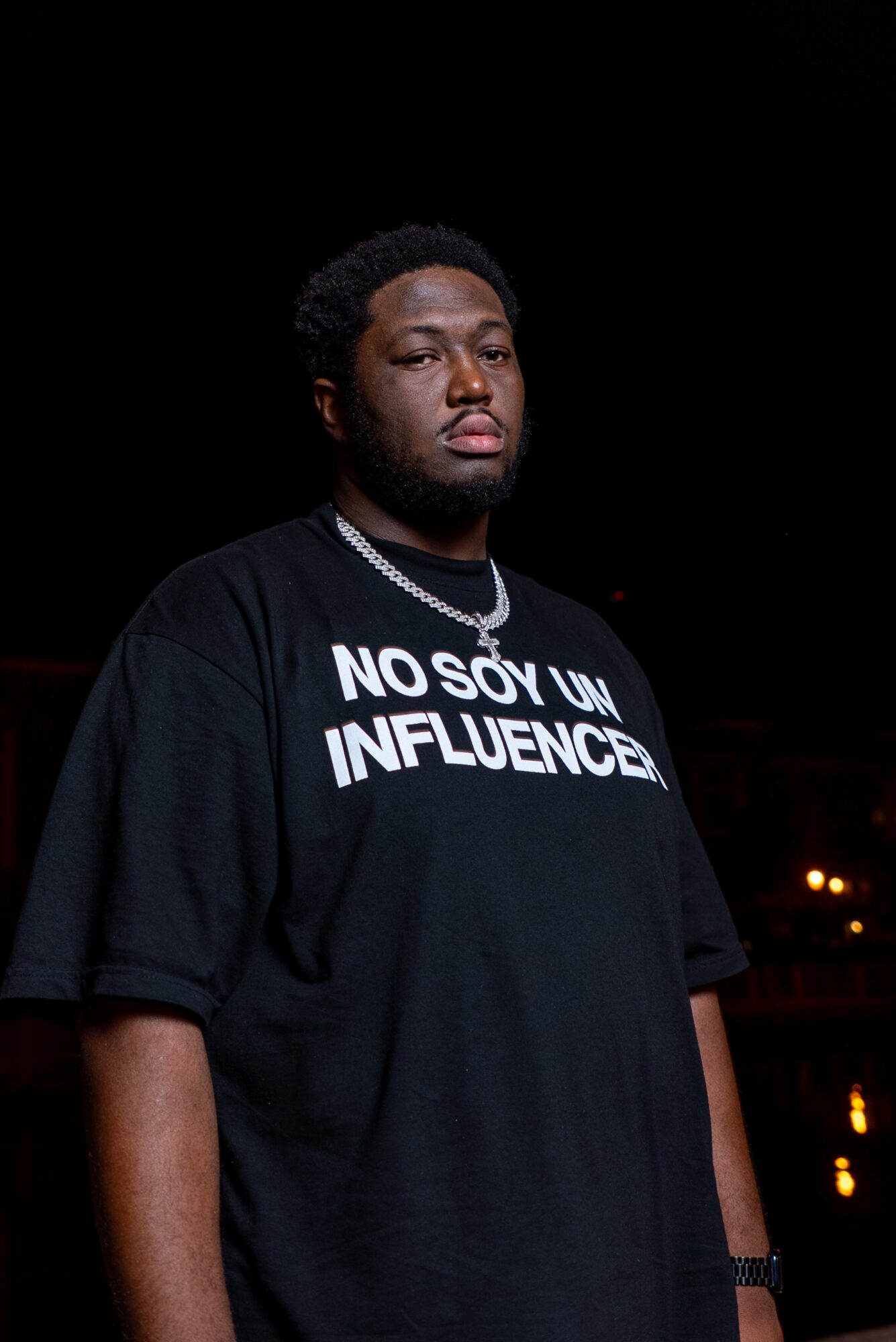 Young man with dark hair and beard wearing a black T-shirt with white text, standing outdoors at night.