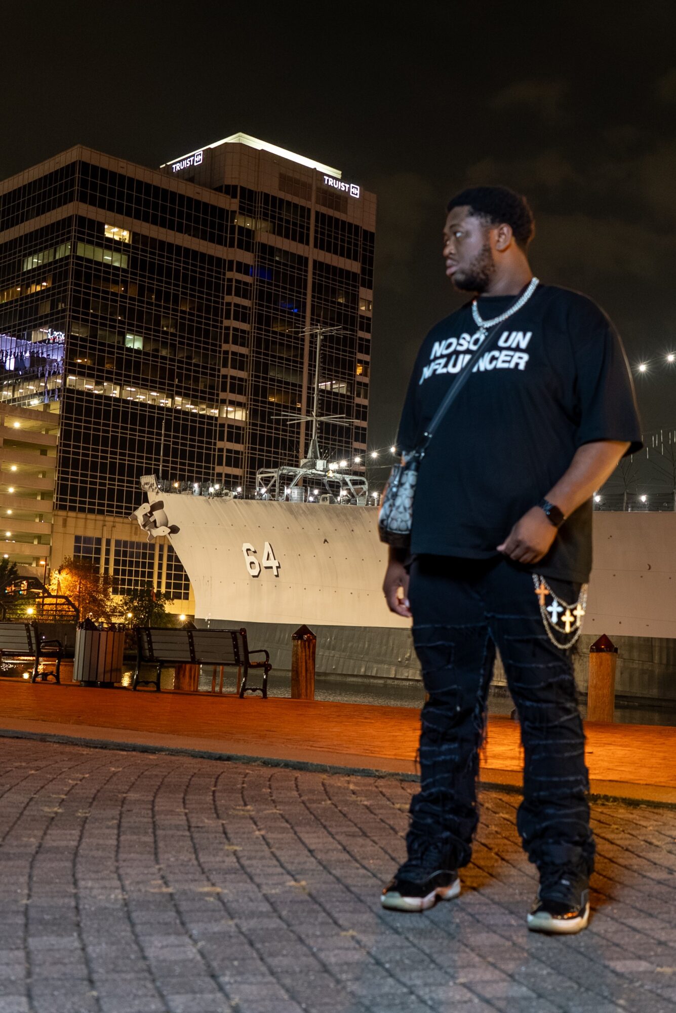 Young man standing outdoors at night with city buildings illuminated behind him, wearing a black T-shirt and jeans.