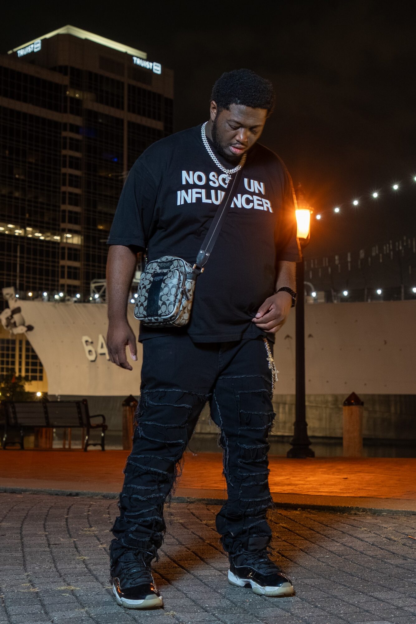 Young man standing outdoors at night, wearing a black hoodie with white text, dark pants, and sneakers, with city buildings in background.