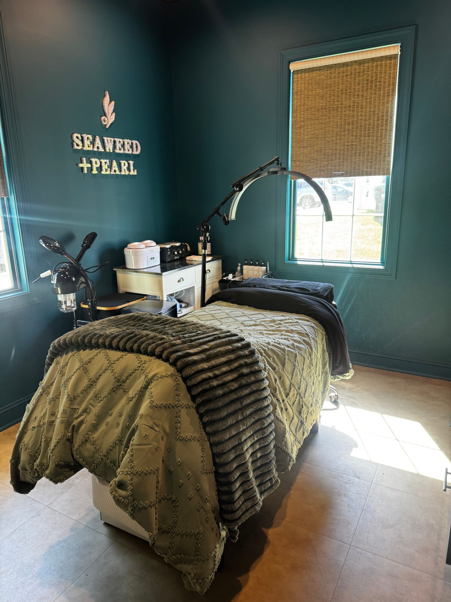 A massage table with a blanket and pillow in a room with teal walls and a window, sunlight on the floor.