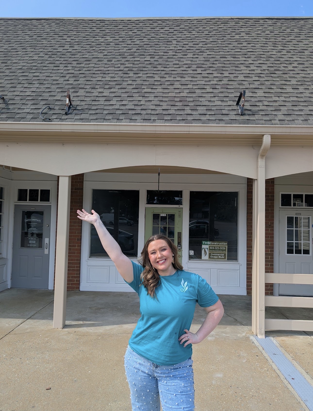 Smiling woman in blue shirt waving outside building with large windows and a porch, under a clear blue sky.