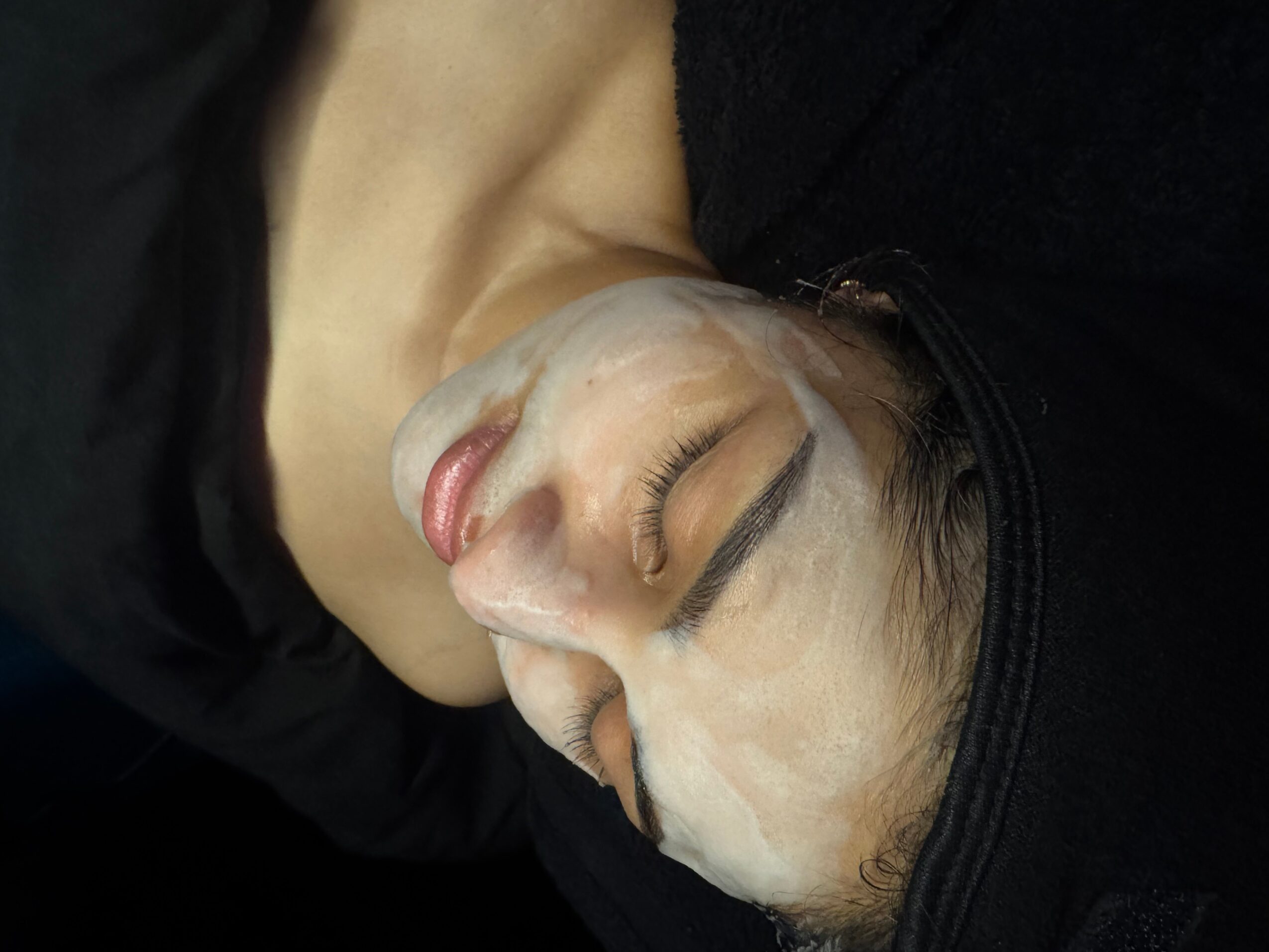 Person lying down with face covered in white cream, eyes closed, wearing a black head covering.
