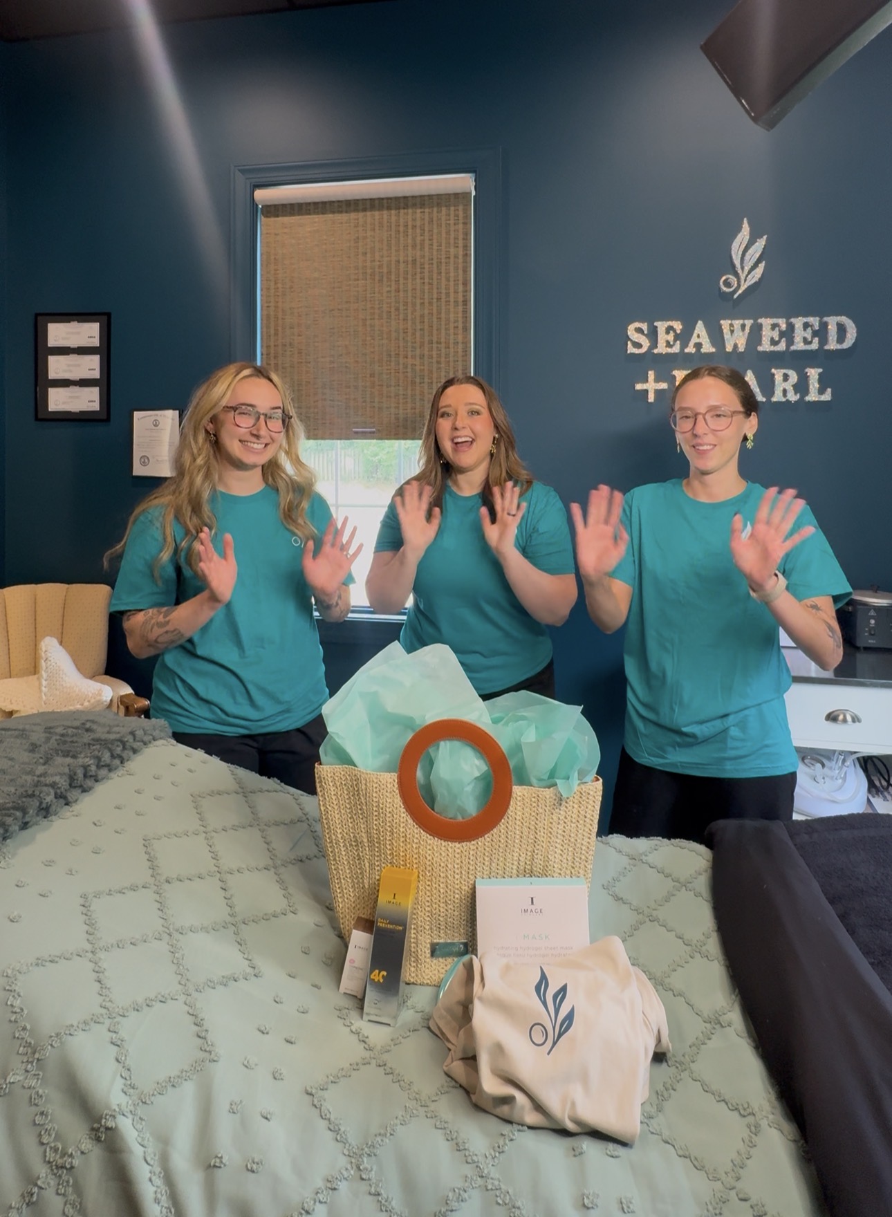 Three people in teal shirts standing behind a table, smiling and waving, in a room with a blue wall and Seaweed + Pearl sign.