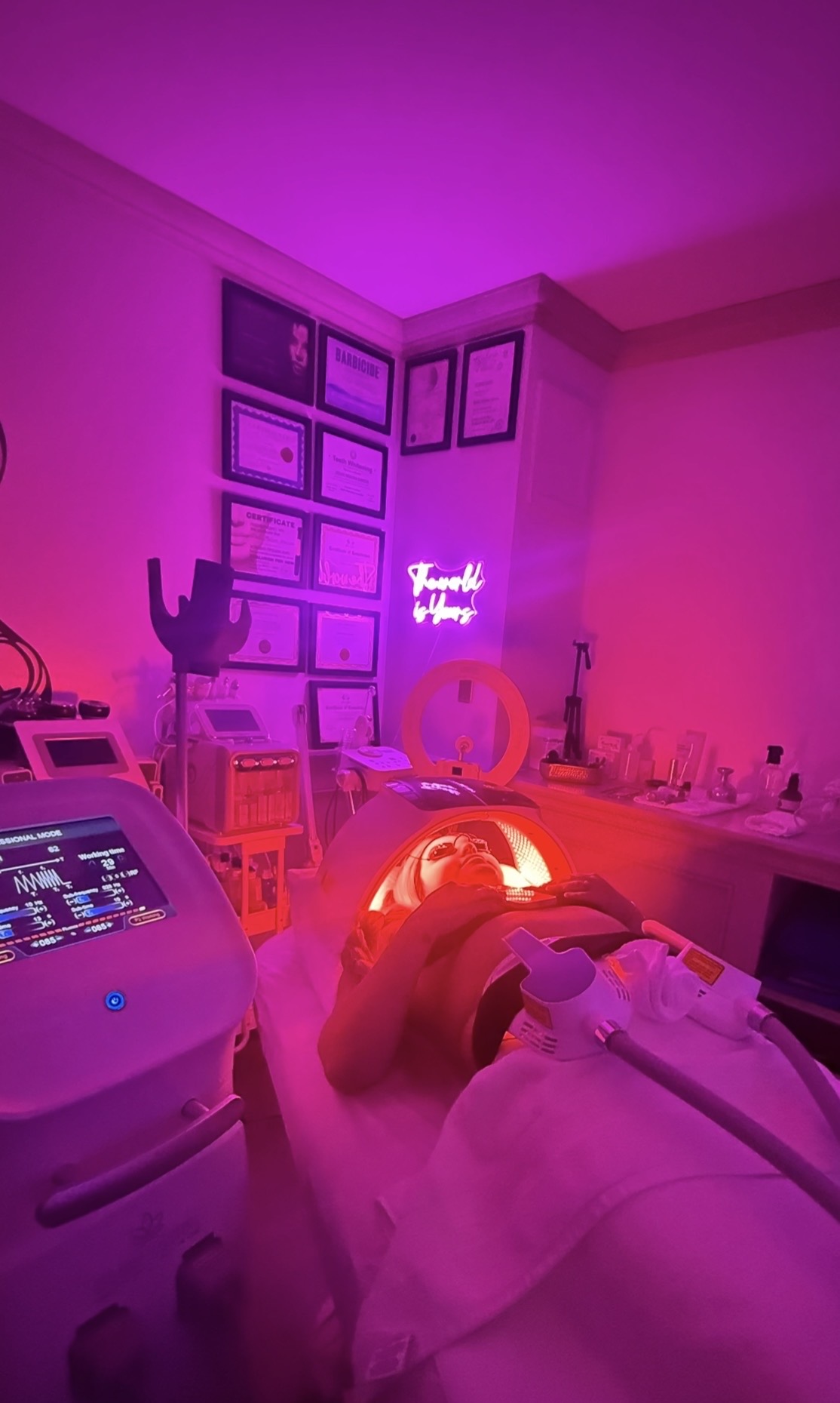 Person receiving a facial treatment in a room illuminated with pink and purple lighting, with skincare equipment and framed pictures on the wall.
