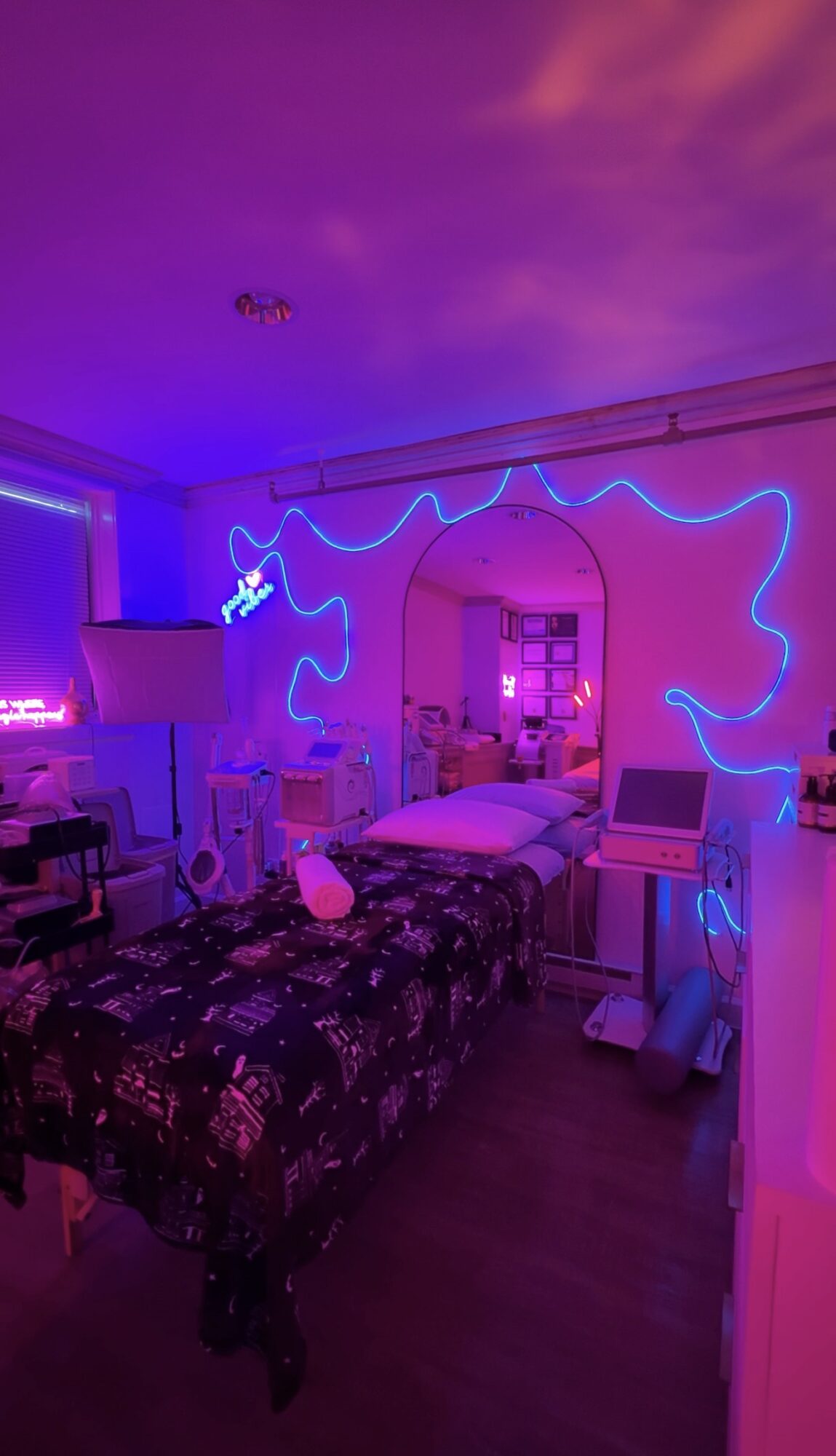 Bedroom with purple and pink lighting, neon blue wall art, bed, and various furniture, creating a vibrant atmosphere.