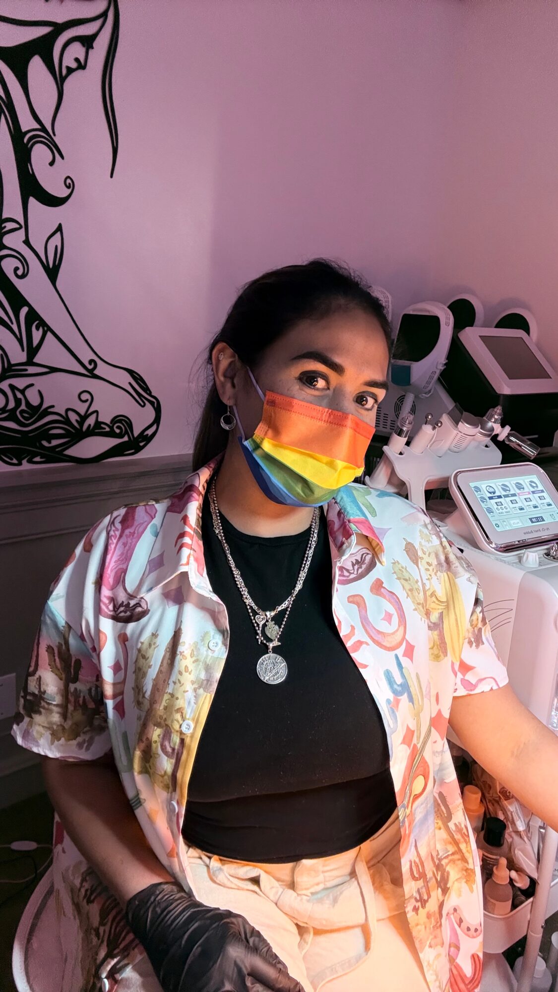 Person wearing a rainbow face mask, black top, colorful jacket, and jewelry, in a pink room with medical equipment.