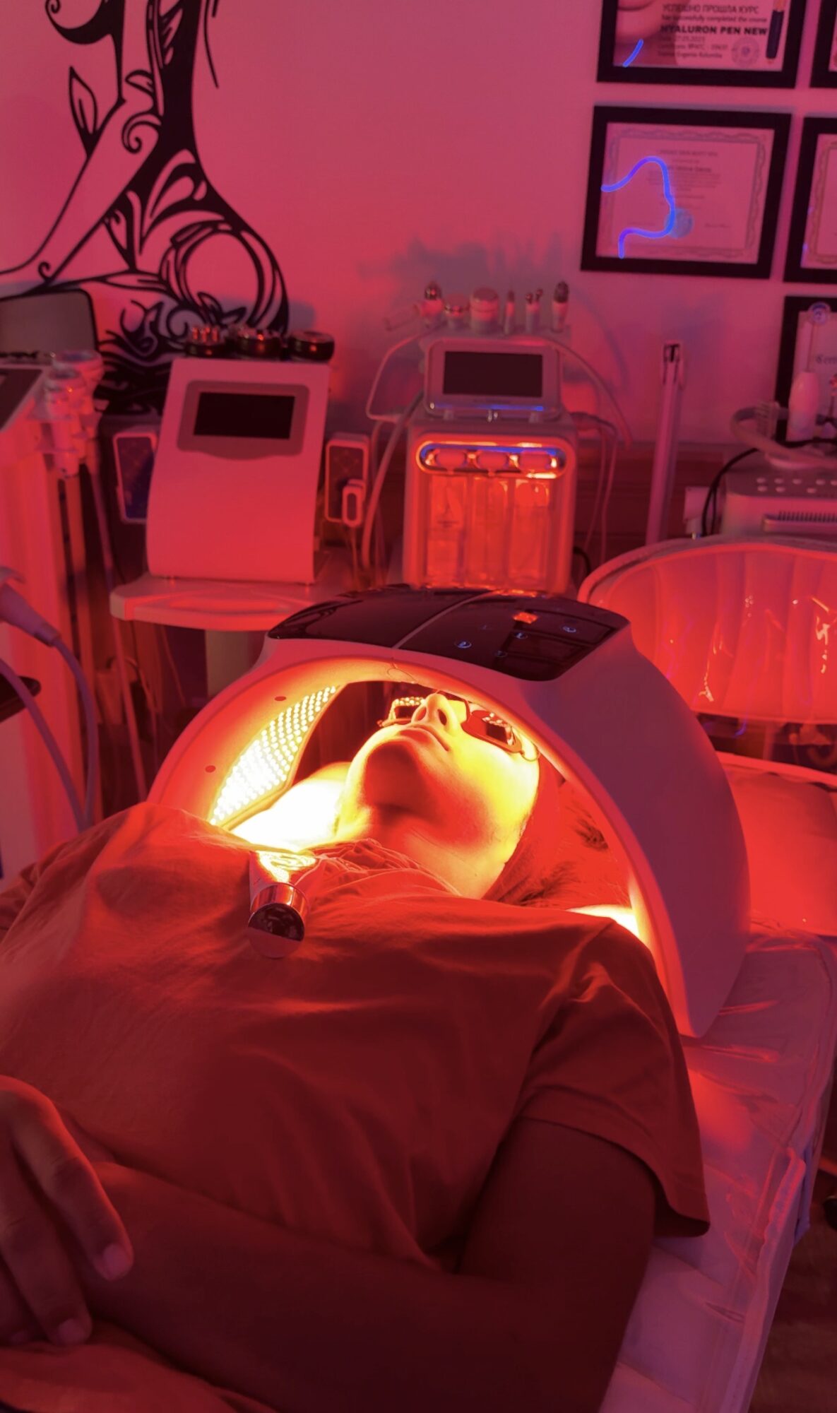 Person lying under a red light in a medical or spa setting, with equipment and framed pictures on the wall behind.