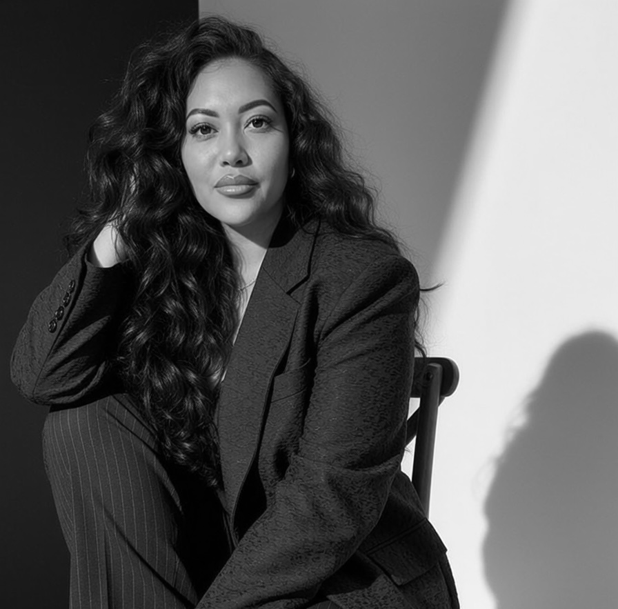 Woman with long curly hair in a blazer, sitting and looking at the camera, shadow on background