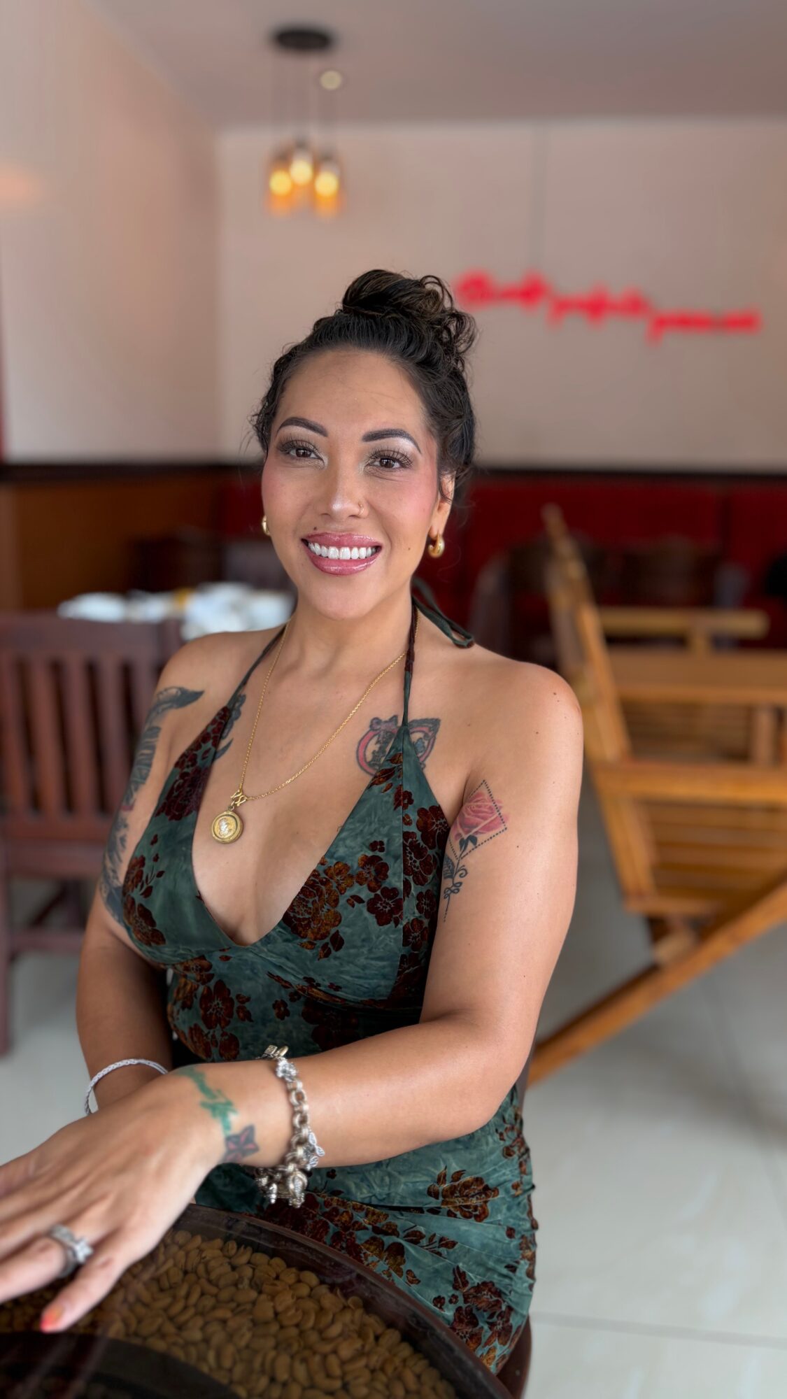 Smiling woman with dark hair in a bun, wearing a patterned dress and jewelry, sitting in a restaurant.