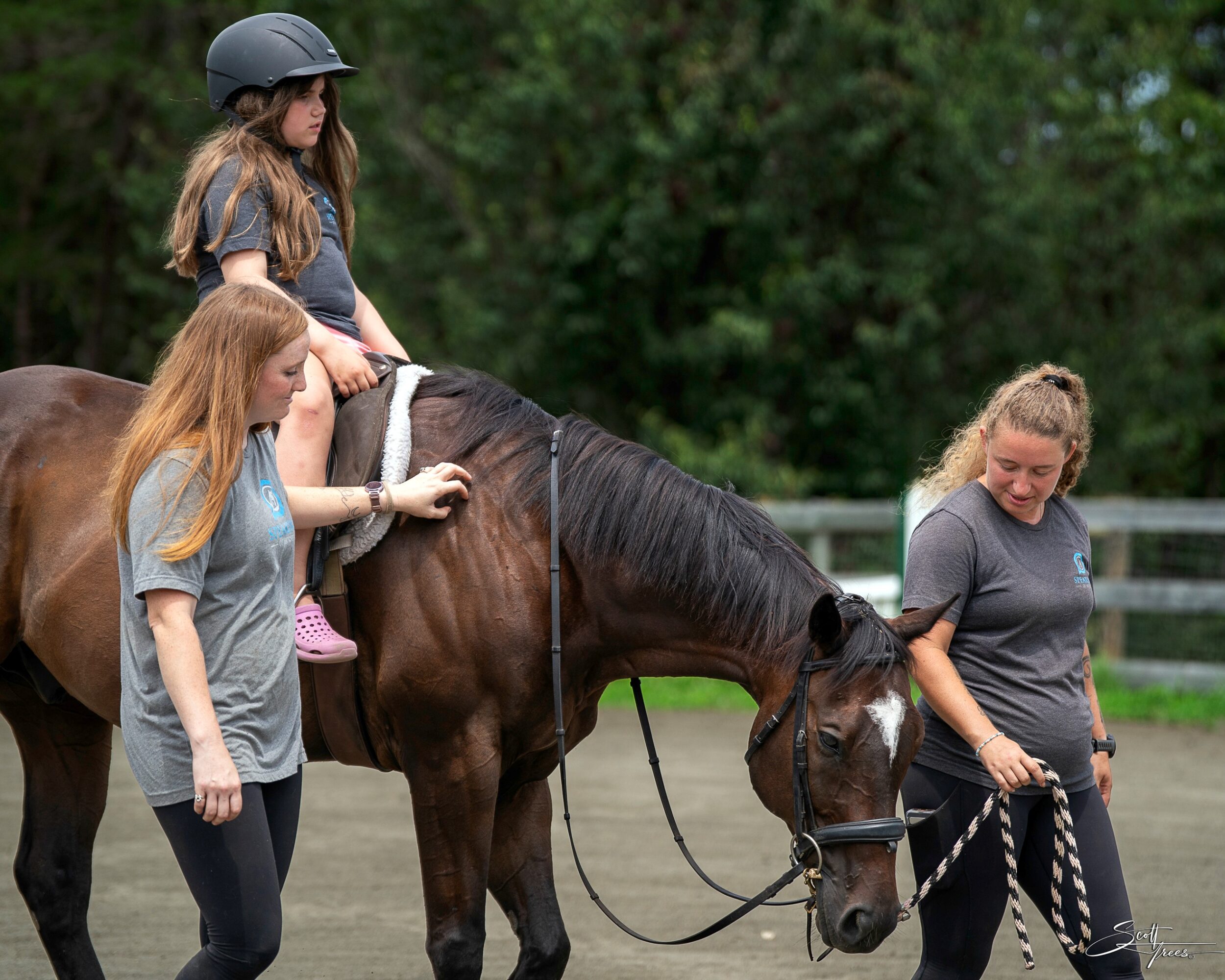 Two women and a girl with a horse outdoors, one girl riding, others guiding the horse, trees in background.