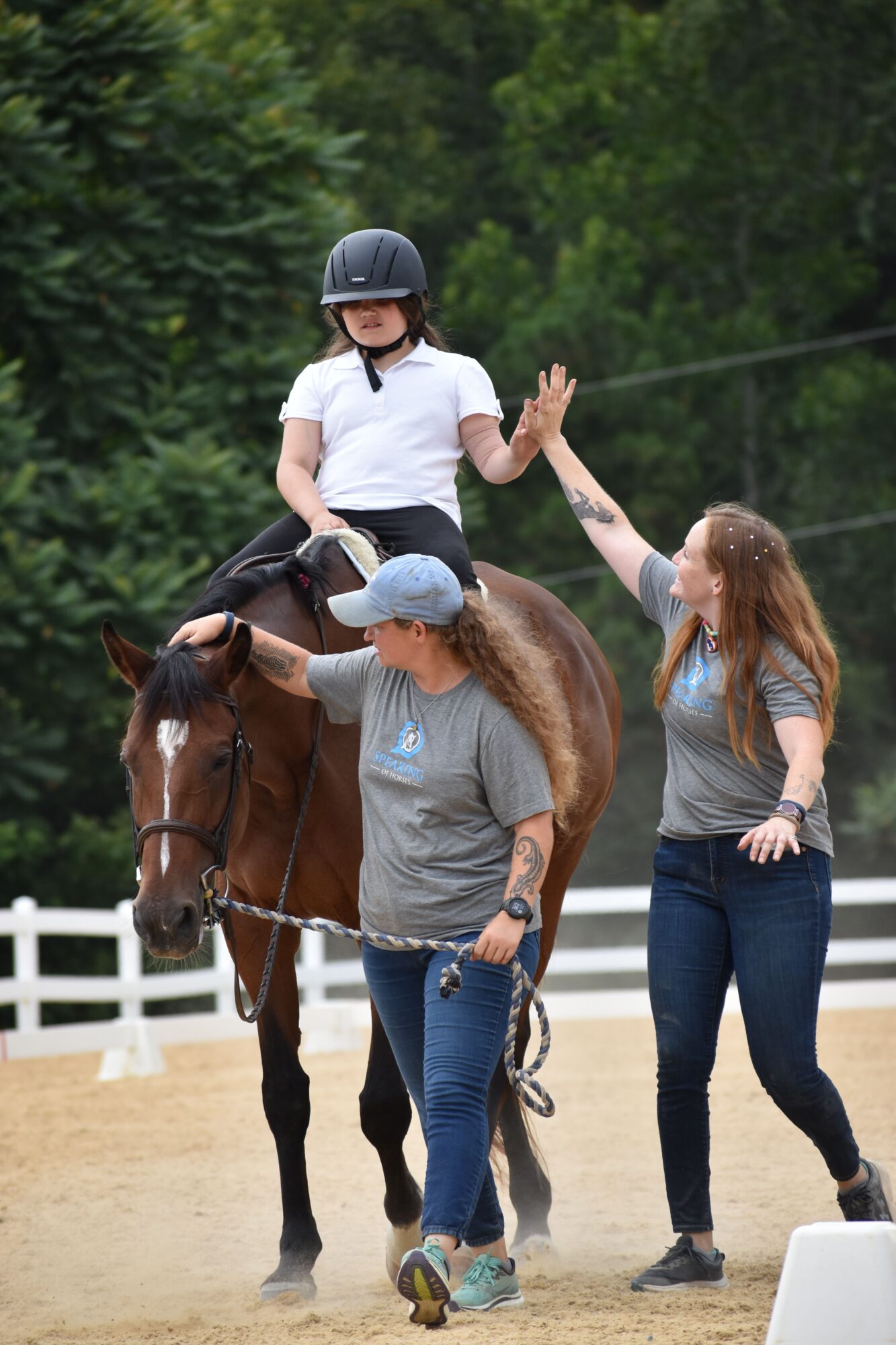 Person riding a horse with a person standing beside, both outdoors near trees and a white fence, during daytime.