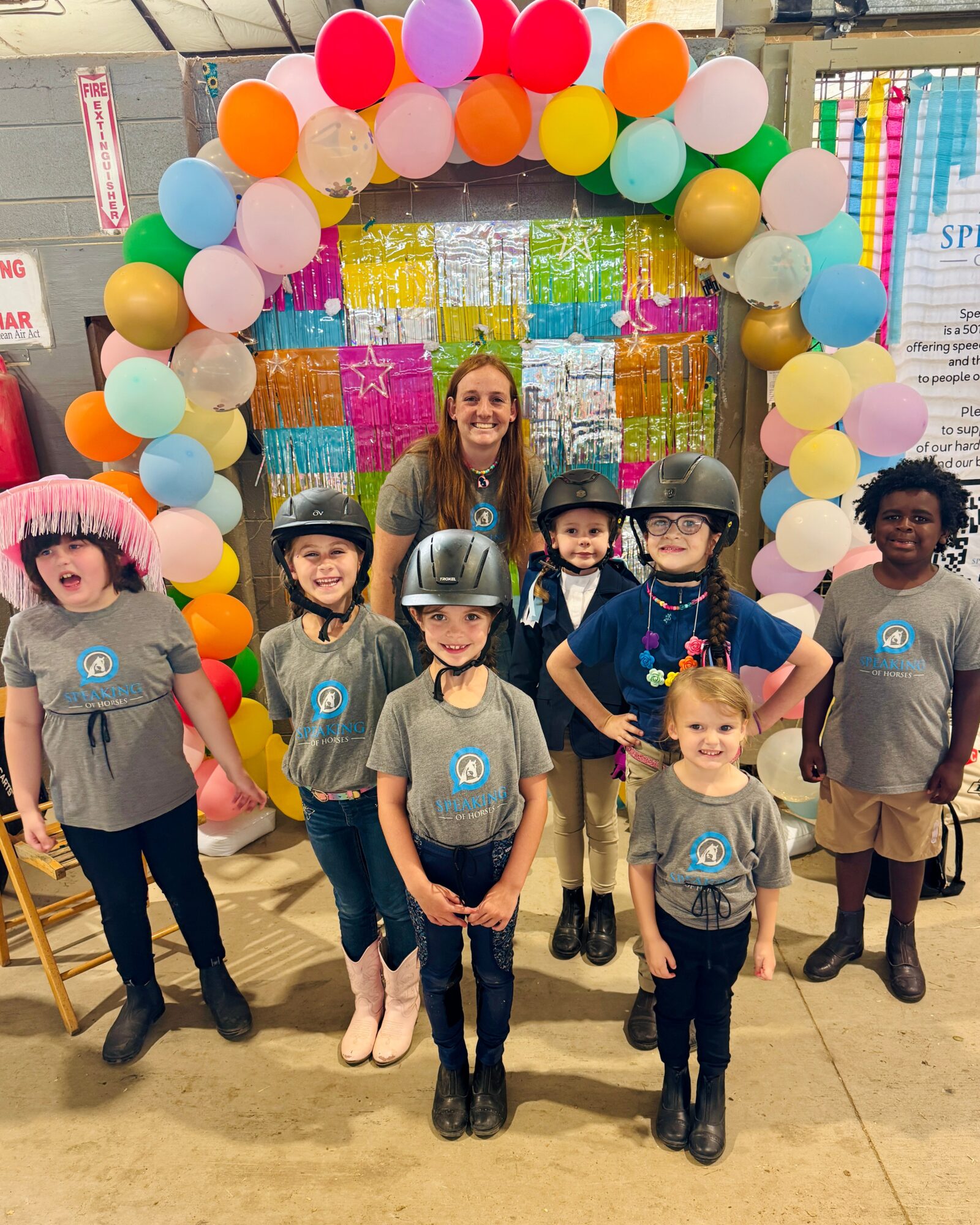 Group of children and an adult standing in front of a colorful balloon arch at an indoor event, smiling.