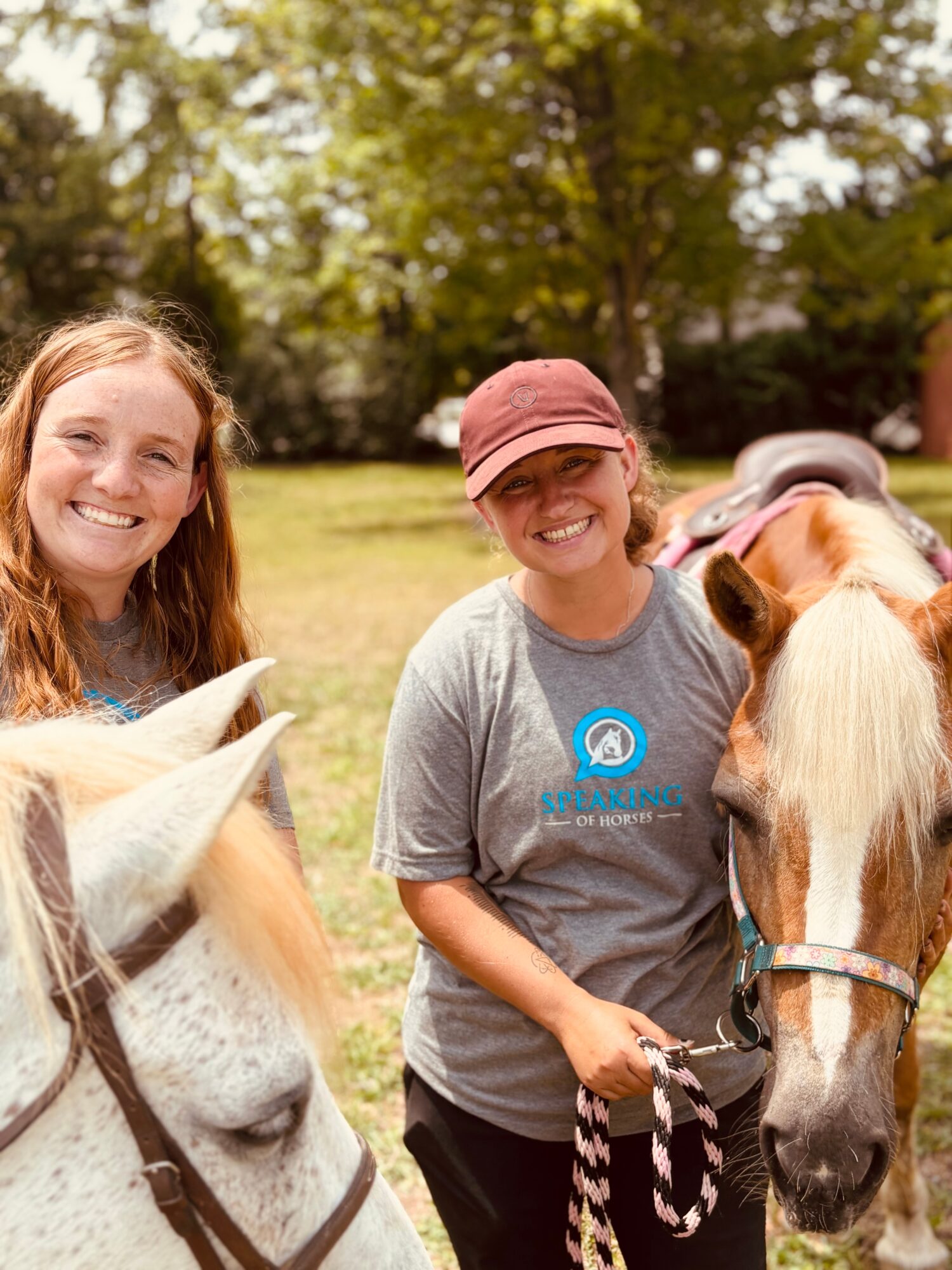 Two smiling women with horses outdoors in a park, trees in background, sunny day.