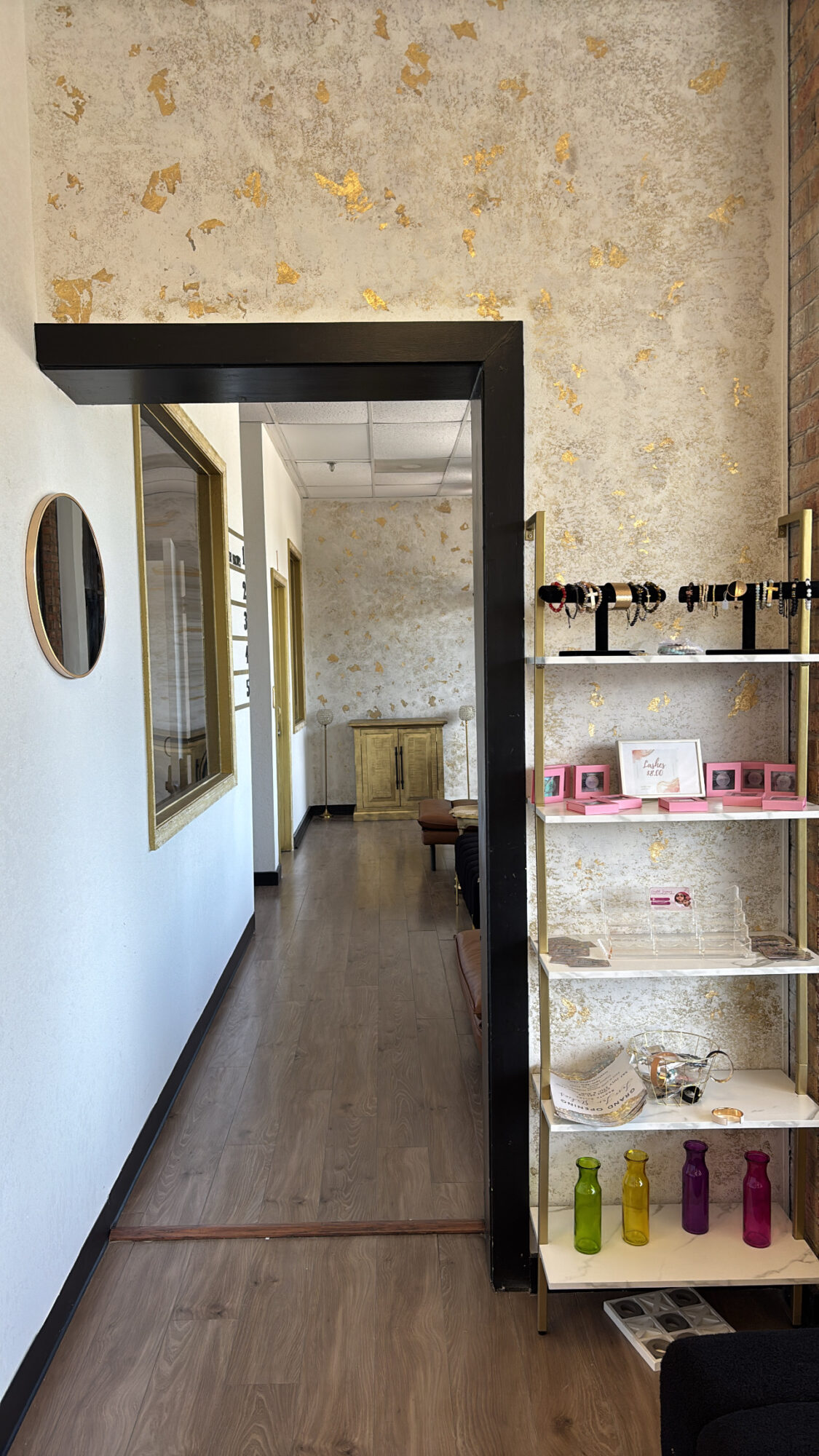 View of a hallway with a doorway, a mirror on the wall, and shelves with bottles and books.